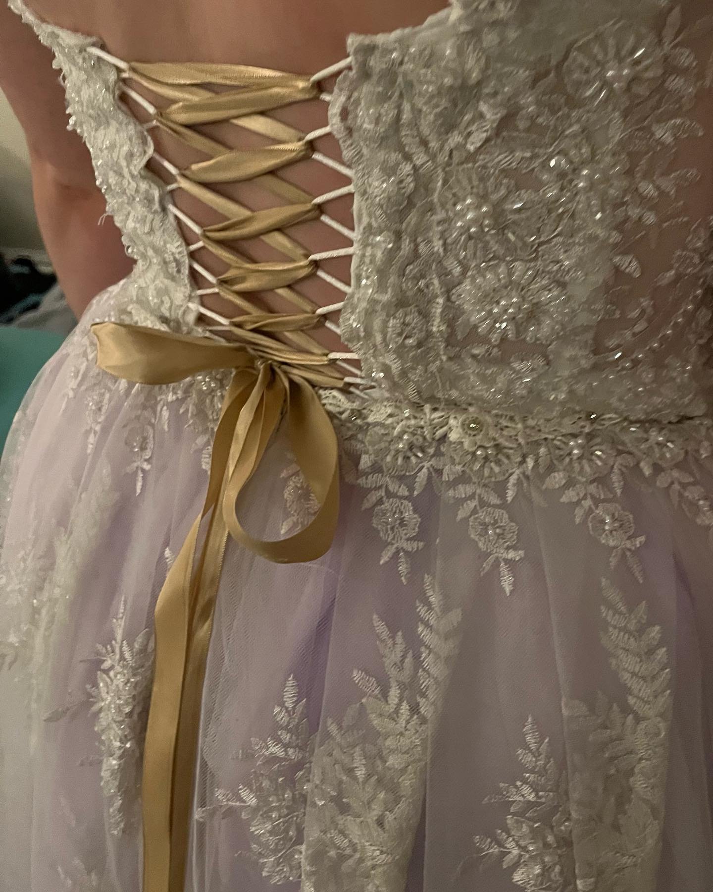 Close-up of a wedding dress with lace and floral embroidery detail, gold ribbon laced through the back, tied into a bow.