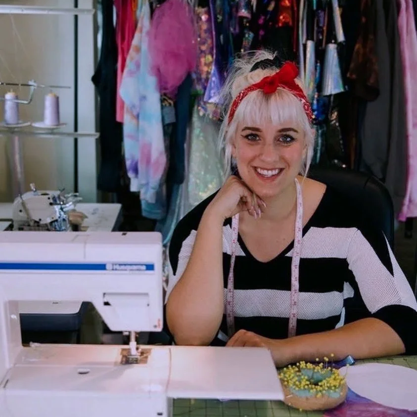 Woman with blond hair and a red bandana, smiling at a sewing machine in a craft room, with colorful fabric and dresses hanging in the background.