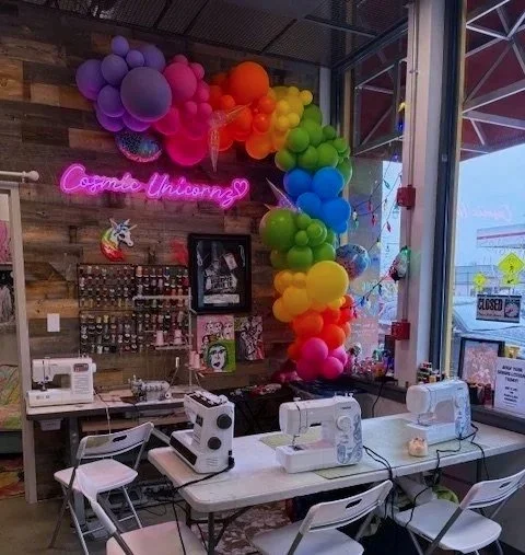 A colorful balloon arch is decorated inside a shop with a neon sign that reads "Cosmic Unicorn". The shop has sewing machines on tables, a wooden wall with a display of small items, and a large window showing the outside streets.