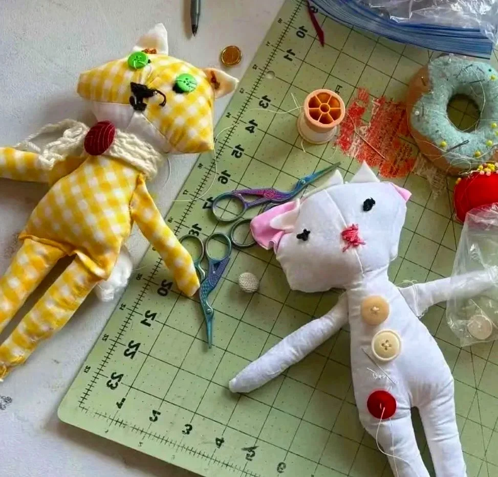 A sewing workspace with two handmade plush dolls, one resembling a yellow checkered cat and the other a white mouse, surrounded by sewing tools including scissors, a needle, thread, pins, buttons, and fabric. The dolls have button and embroidered facial features and are laid on a cutting mat with a measuring grid.