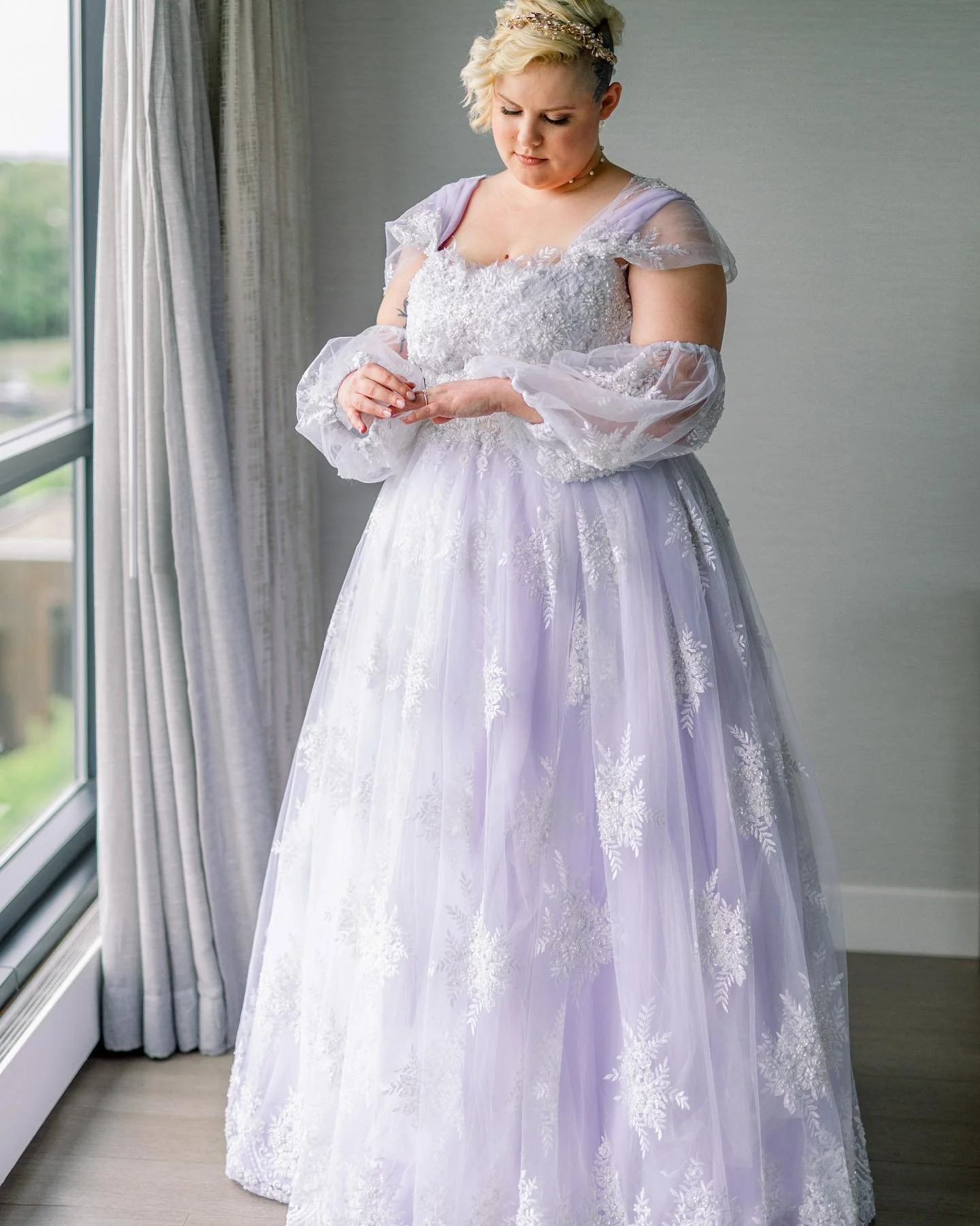A woman in a white, ornate wedding dress with sheer sleeves and lace details, standing by a window inside a room.