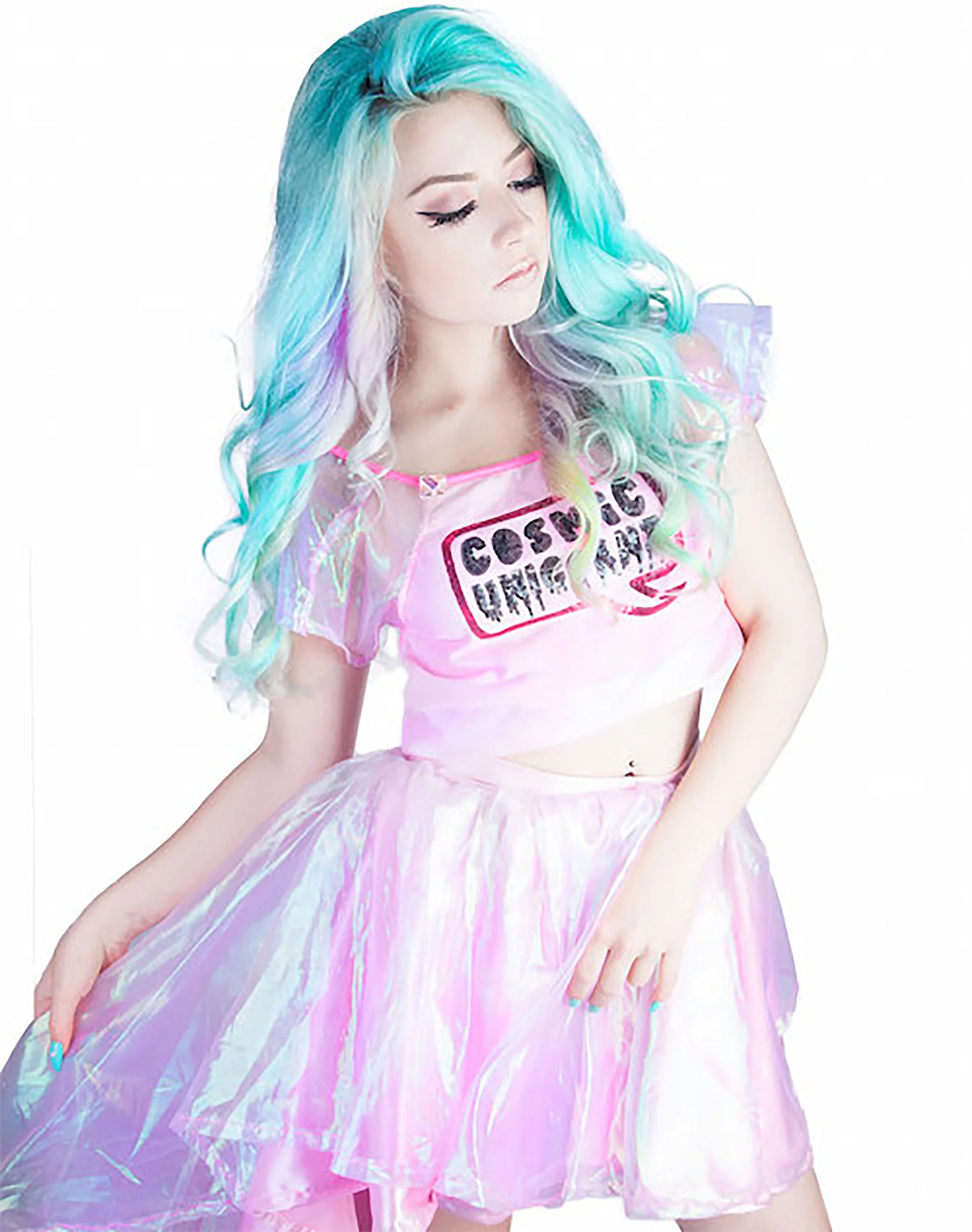 Young woman with pastel blue, purple, and pink wavy hair wearing a pink iridescent skirt and a pink cropped top with the text 'Cosmic Unisex'. She is looking down with soft makeup against a white background.