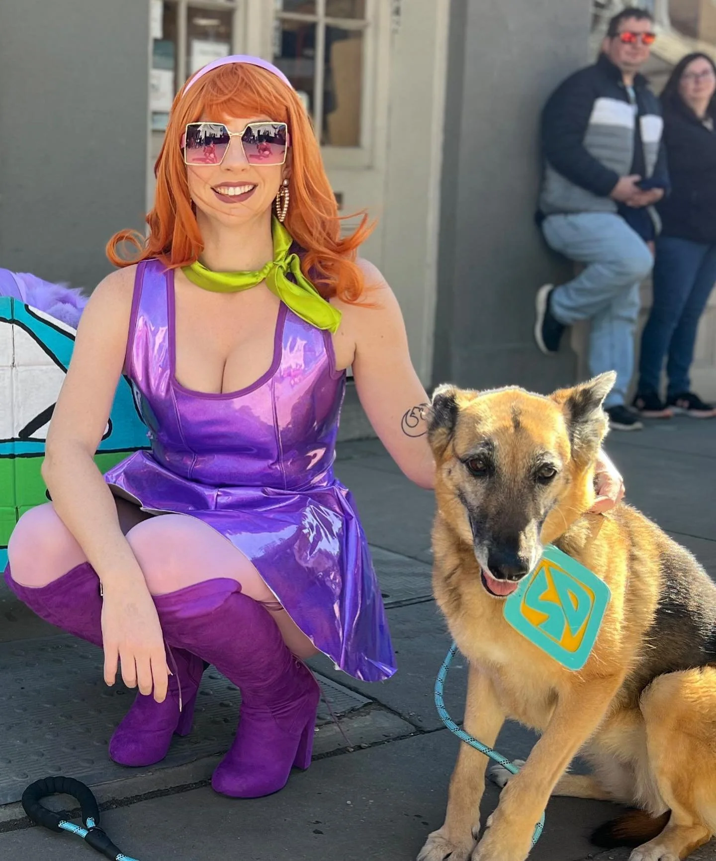 A woman dressed in a colorful costume with purple dress, matching boots, and sunglasses, squatting next to a police dog with a superman emblem on its collar, in an outdoor urban setting with two people in the background.