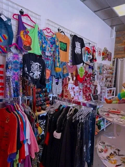 A retail display with colorful clothing hanging on racks and a wall, including t-shirts, dresses, and accessories. There are also gift items and jewelry on tables and shelves.