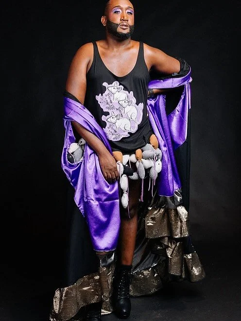 A person with a beard and makeup wearing a black tank top with a cartoon unicorn design, purple satin robe, and high-heeled boots, posing against a black background.