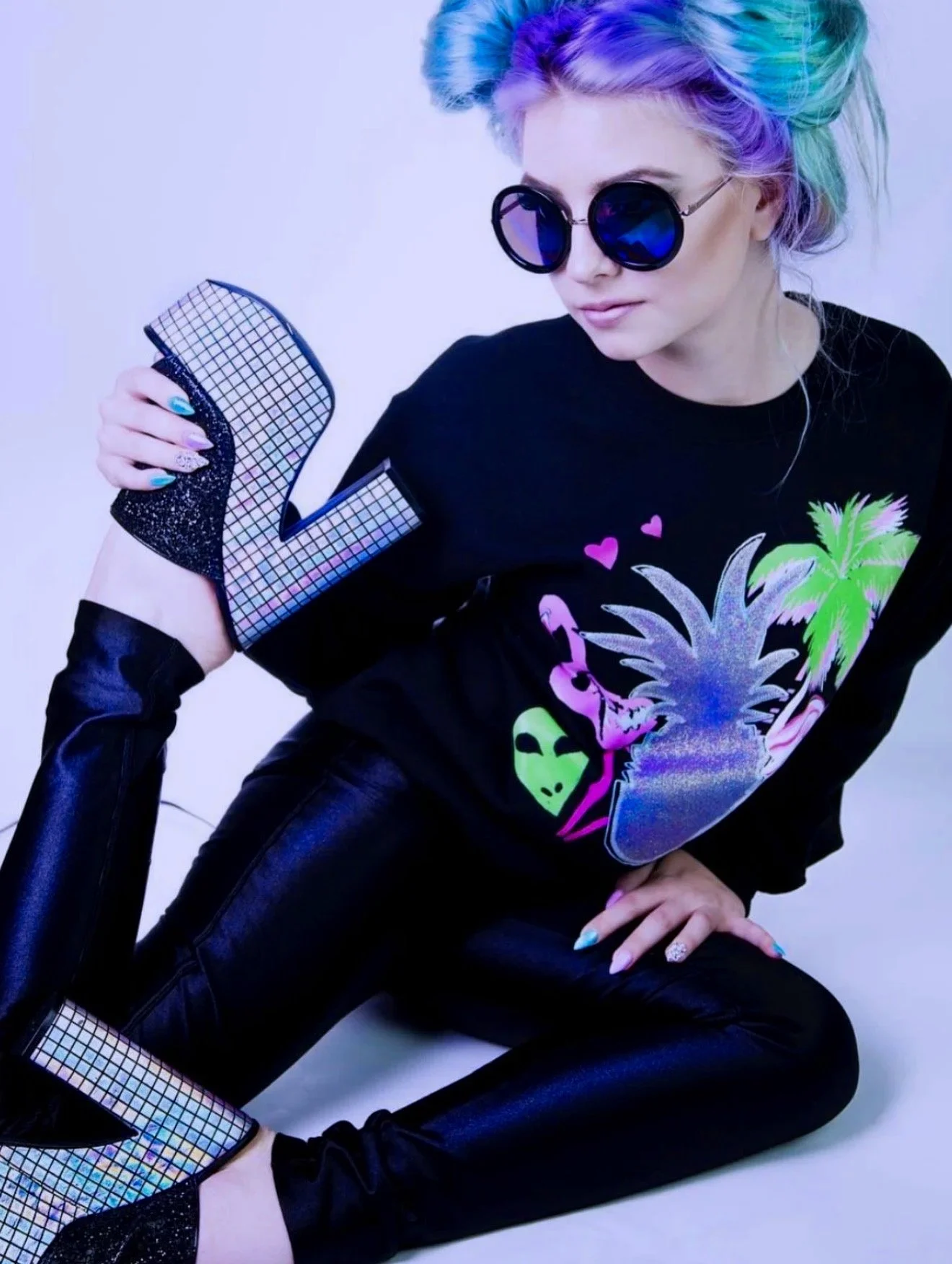 A woman with rainbow-colored hair, wearing black sunglasses, a black graphic t-shirt with neon palm trees and hearts, and shiny, holographic high-heeled boots.