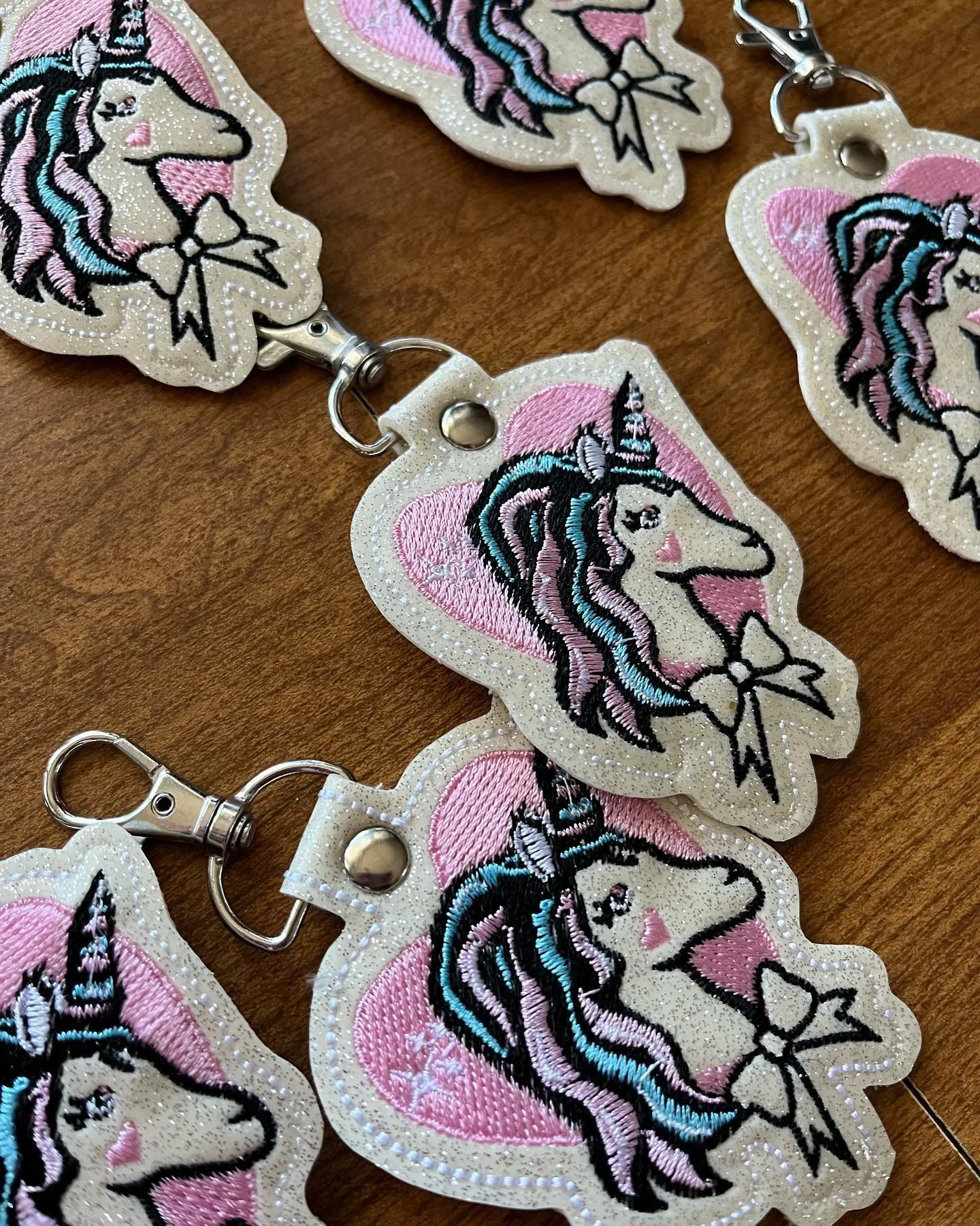 Multiple embroidered keychains featuring a unicorn with a pink background and a bow, attached to metal clasps, on a wooden surface.