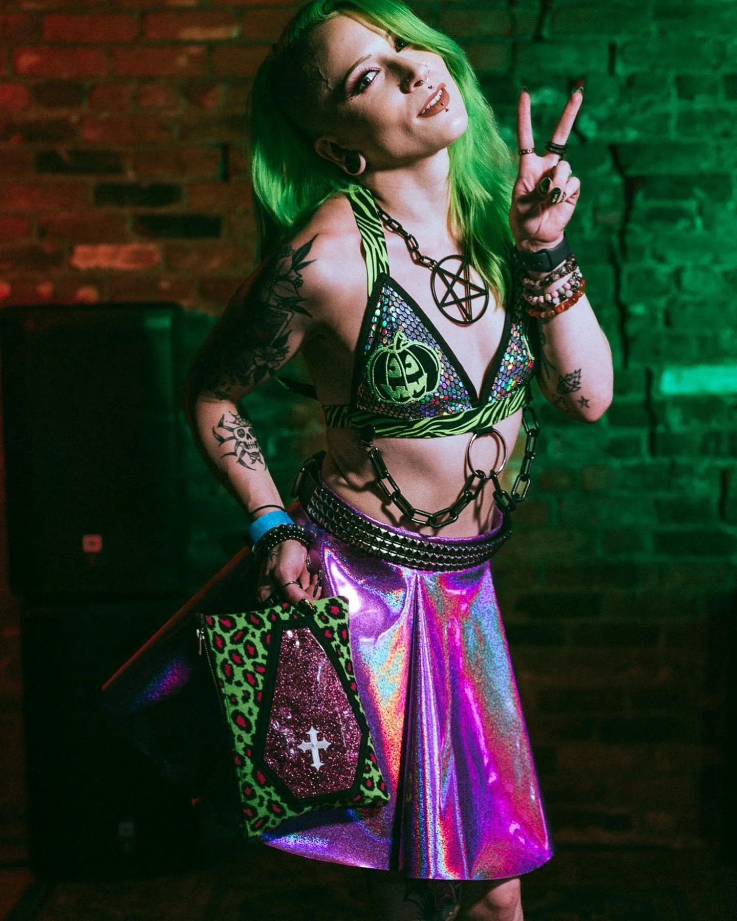 A young woman with bright green hair making a peace sign at a party or nightclub, dressed in colorful, gothic-inspired clothing with tattoos, jewelry, and a holographic skirt, standing against a brick wall with green lighting.