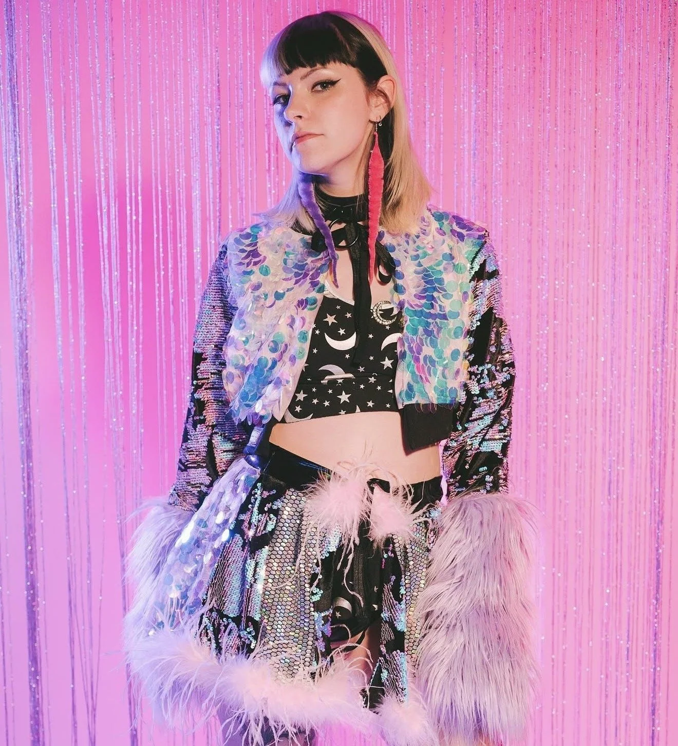 Young woman wearing a colorful sequined jacket, black star and moon crop top, feathered accessories, and a skirt with matching textures, standing against a pink shimmering curtain.