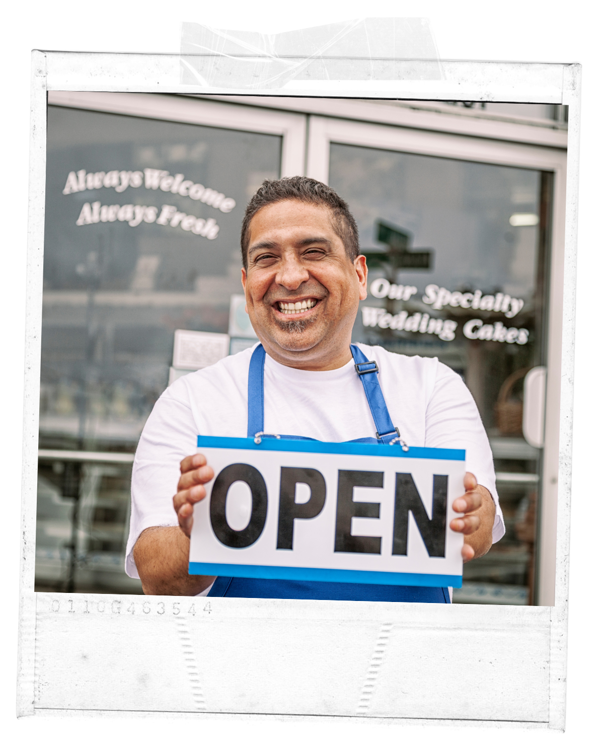 Smiling man wearing an apron holding a sign that says 'OPEN' in front of a storefront with signs that say 'Always Welcome Always Fresh' and 'Our Specialty Wedding Cakes'.