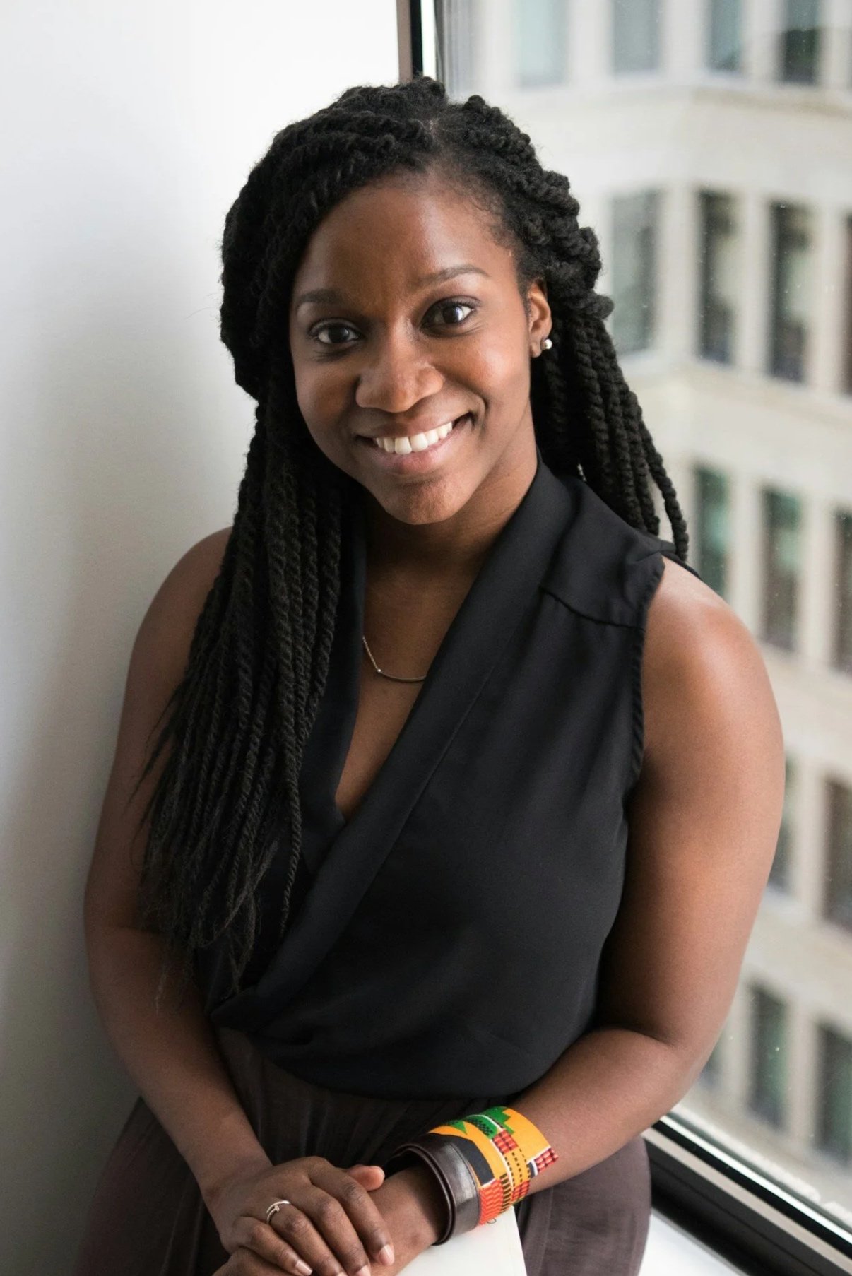 A smiling woman with long braided hair stands near a window with a cityscape view in the background.