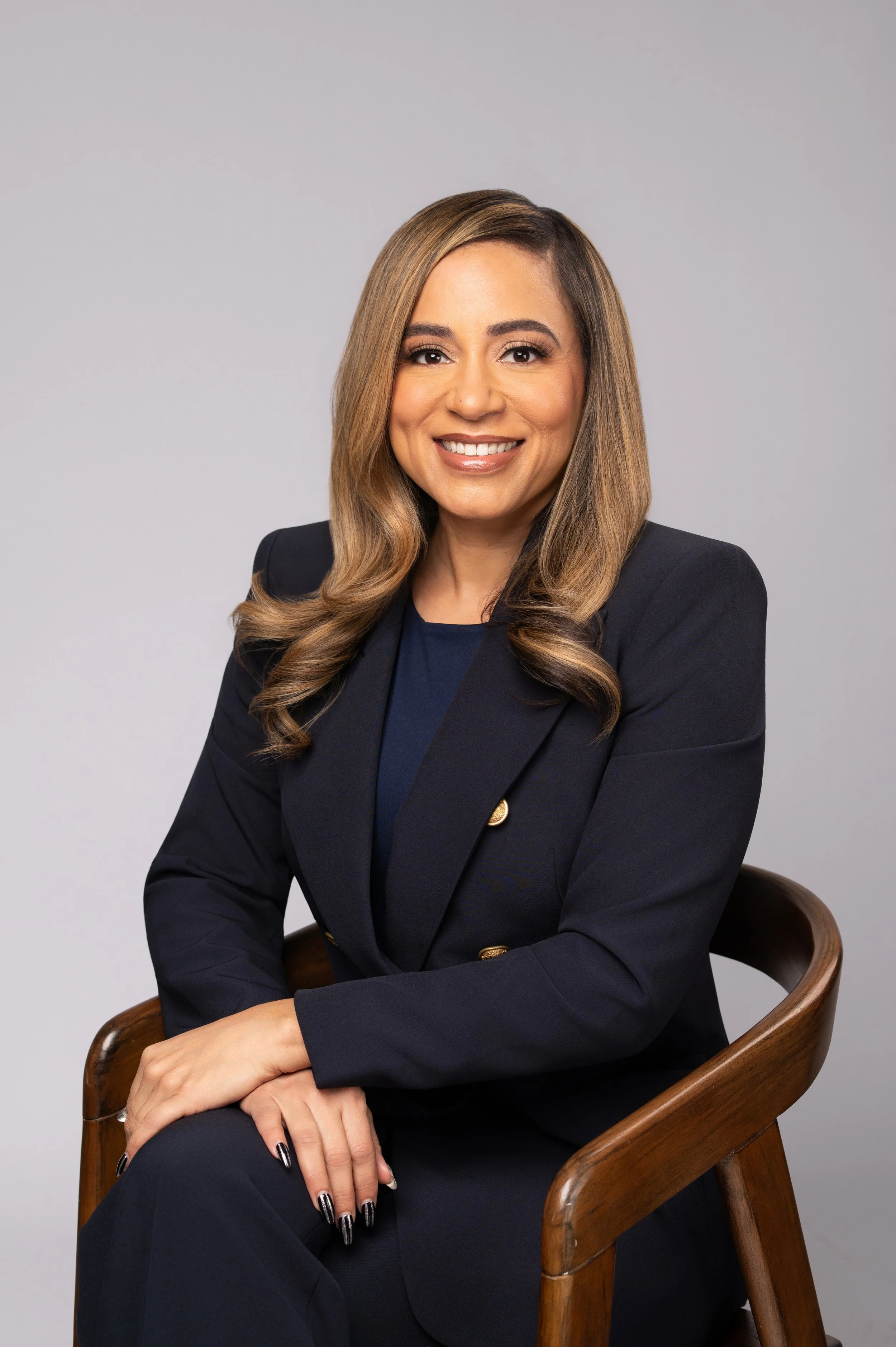 A woman with long, wavy light brown hair and a bright smile, wearing a dark navy blazer with gold buttons and a matching top, sitting on a wooden chair against a plain light gray background.