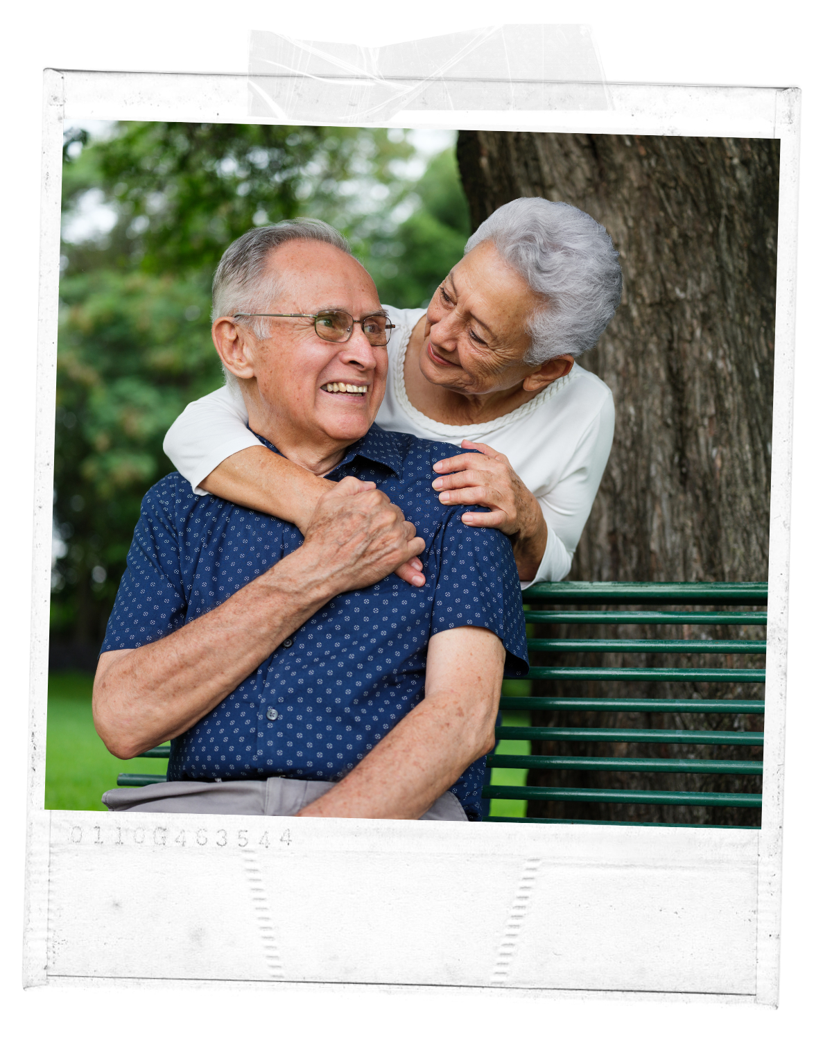 An elderly man and woman sharing a joyful moment outdoors under a tree, sitting on a bench, with the woman hugging the man from behind.