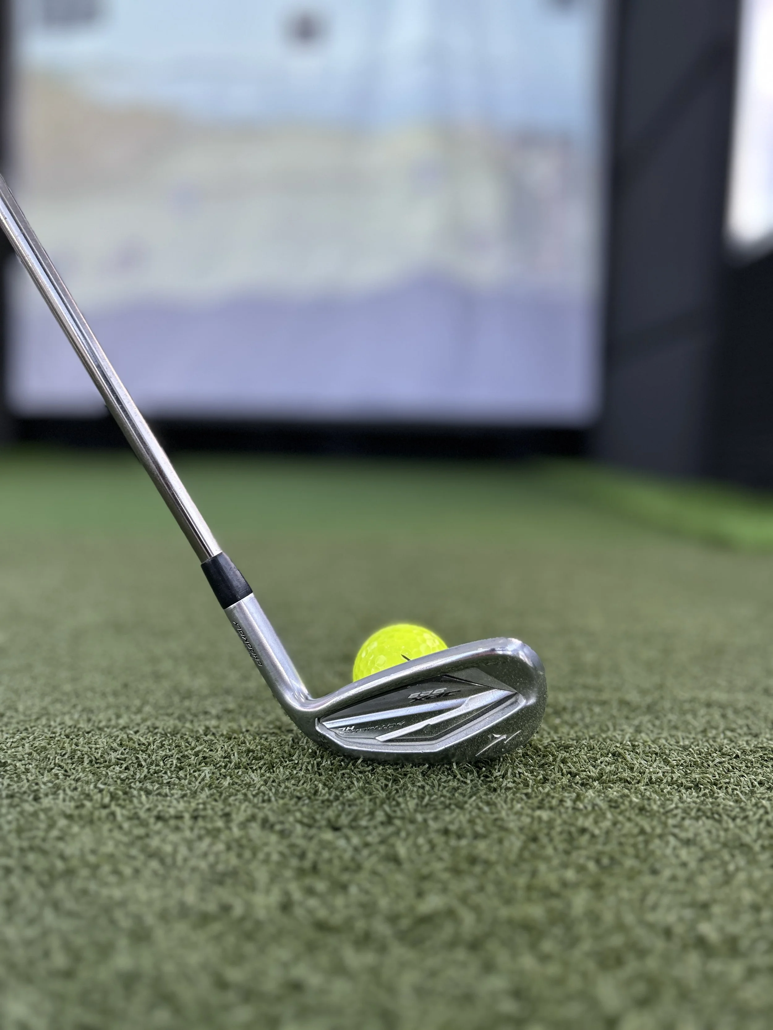 Close-up of a golf club and a yellow golf ball on a green putting surface, with a blurred screen in the background.