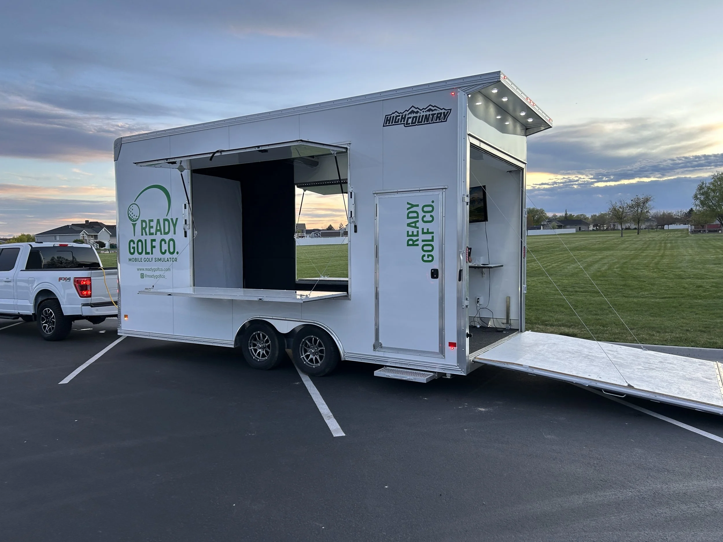 Mobile golf simulator trailer with open side and logo 'Ready Golf Co.' parked on a paved lot at sunset.