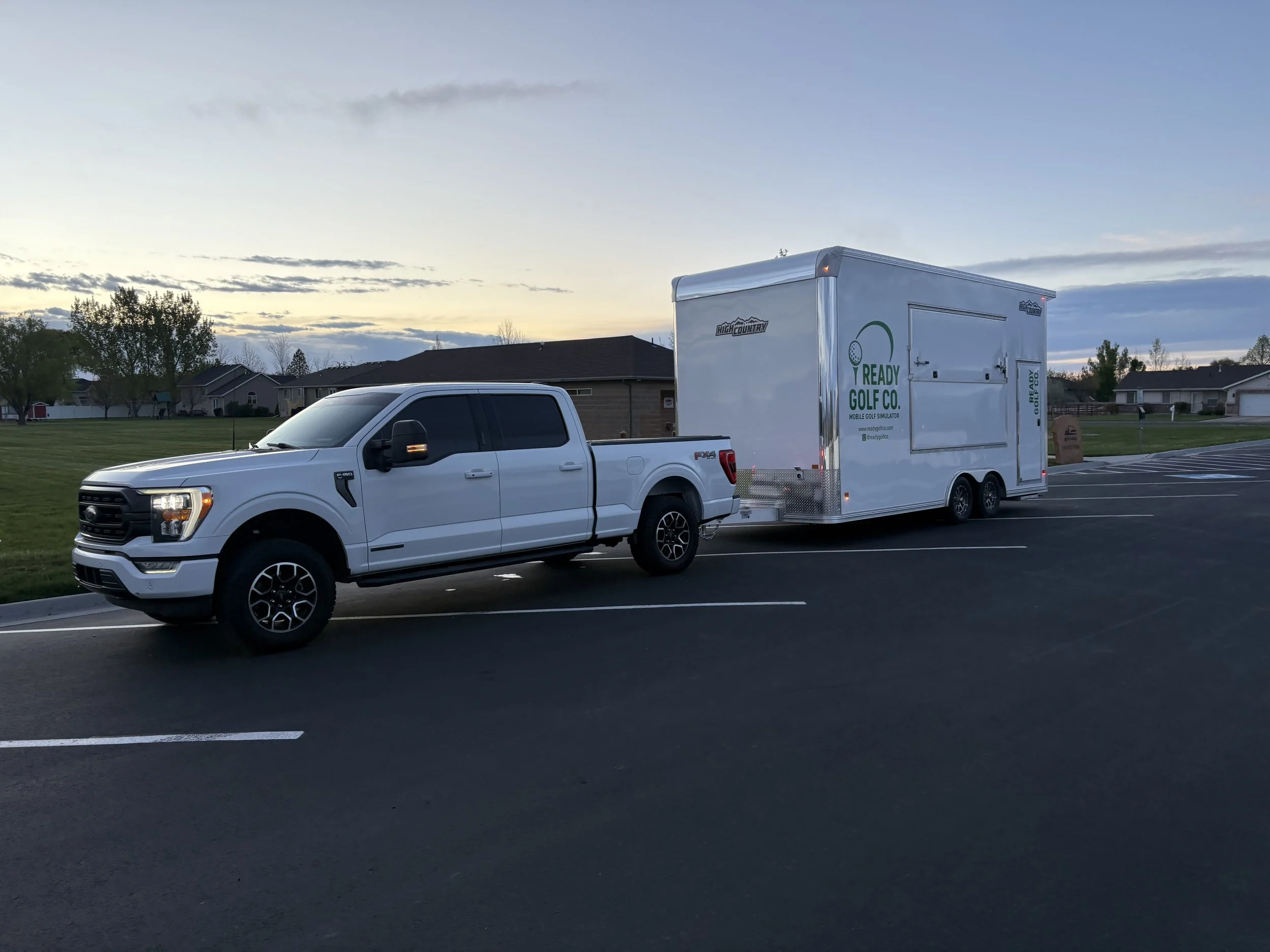 A white pickup truck towing a white trailer with green signage in a parking lot at sunset with houses in the background.