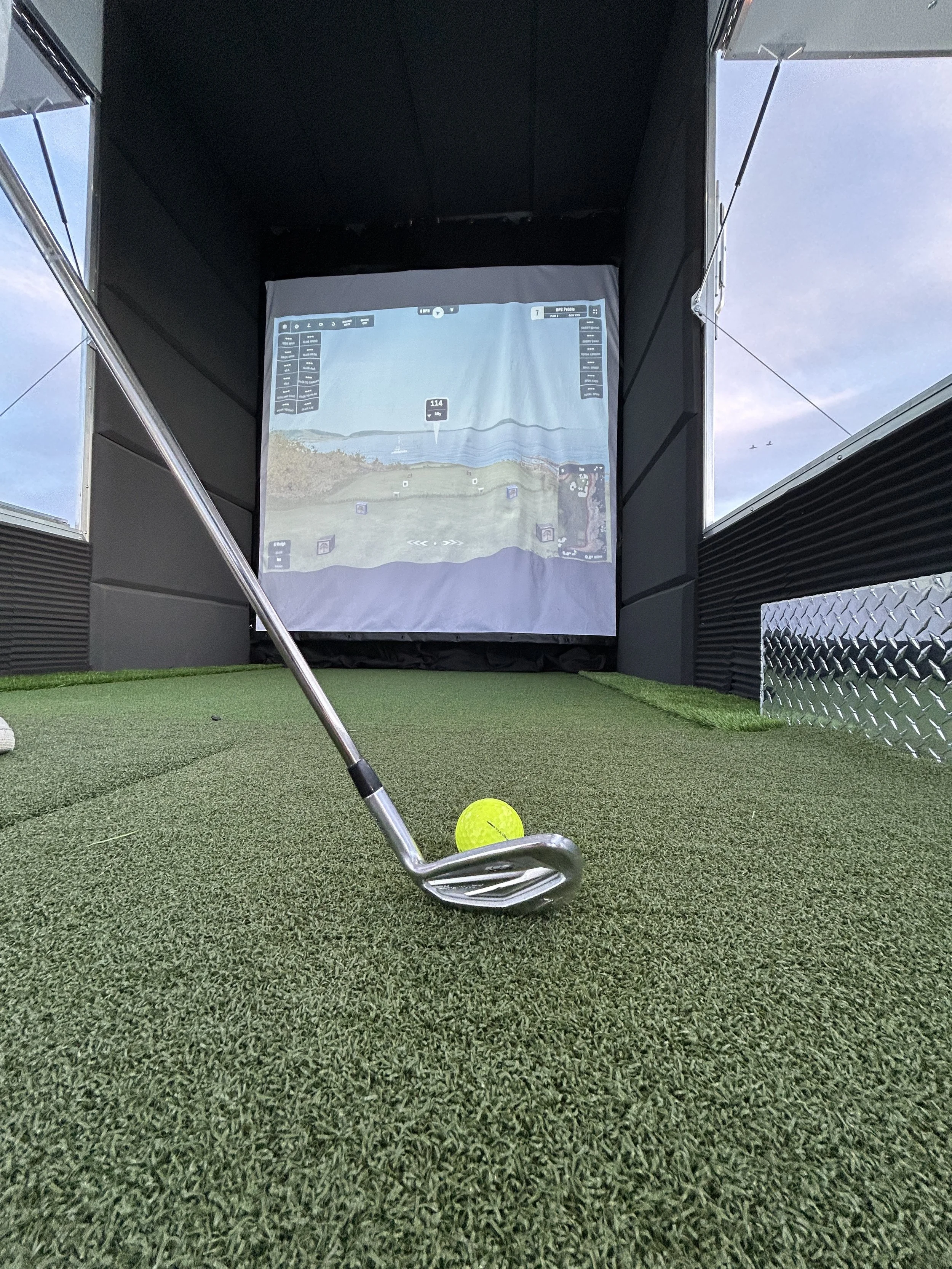 Golf simulator room with a golf club and ball on the artificial turf, and a projected golf course on a screen in the background.
