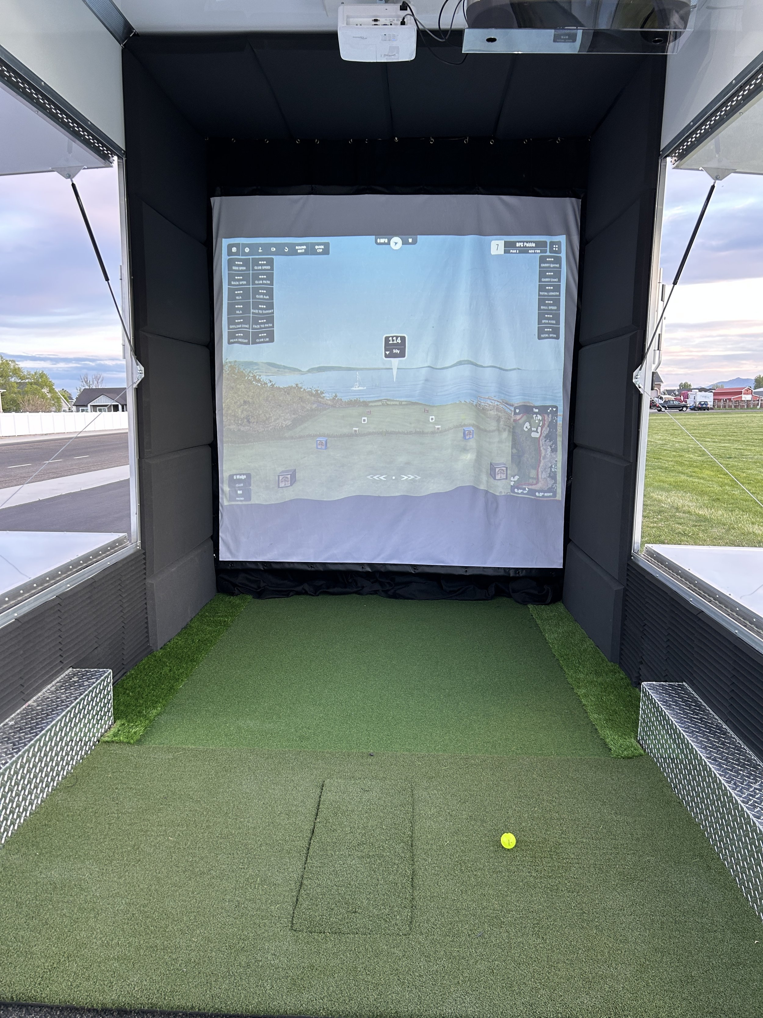 Indoor golf simulator with a virtual driving range displayed on the screen, artificial turf on the floor, and a yellow golf ball resting on the turf.