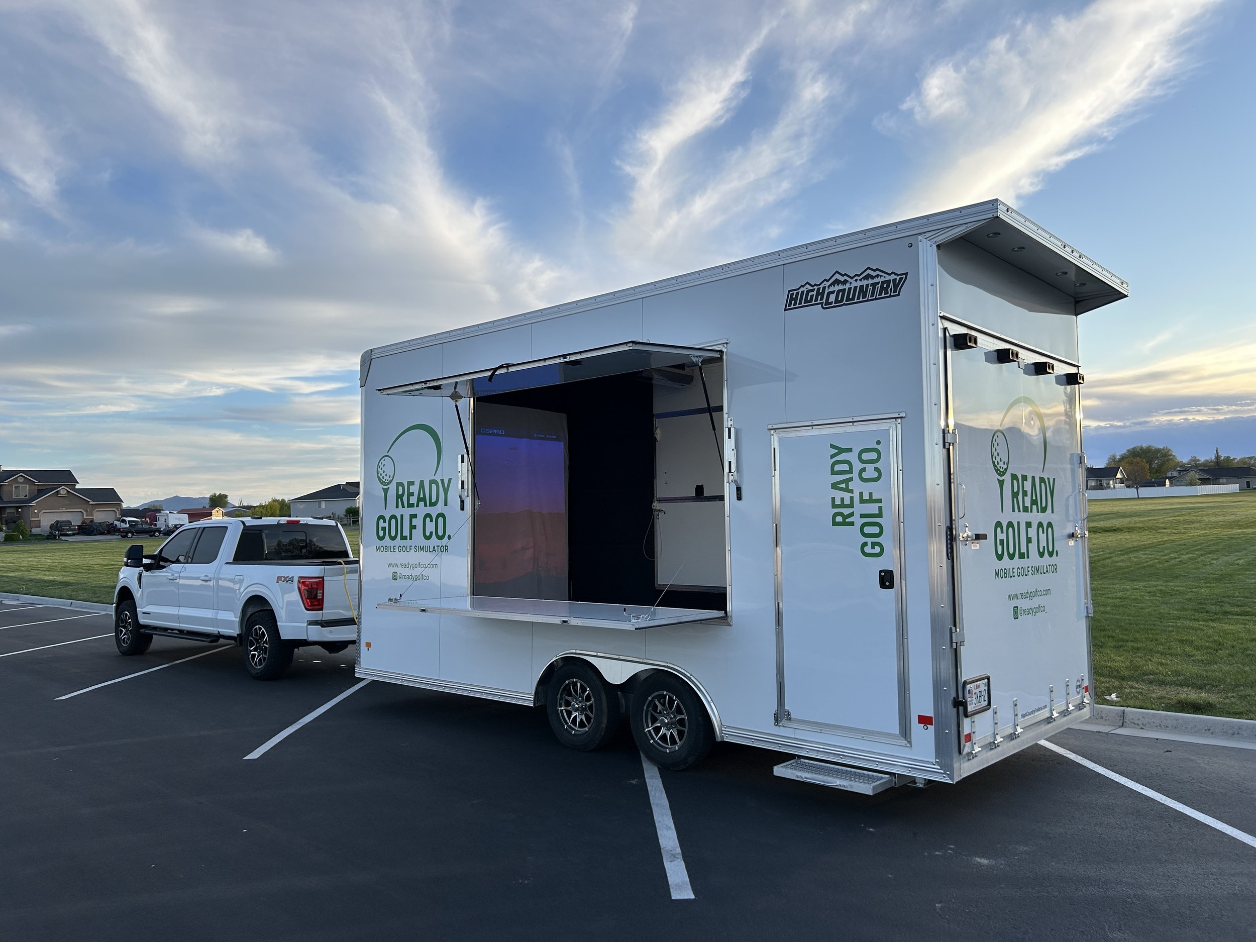 A mobile golf simulator trailer labeled 'Ready Golf Co.' parked in a parking lot next to a white pickup truck against a sky with some clouds.