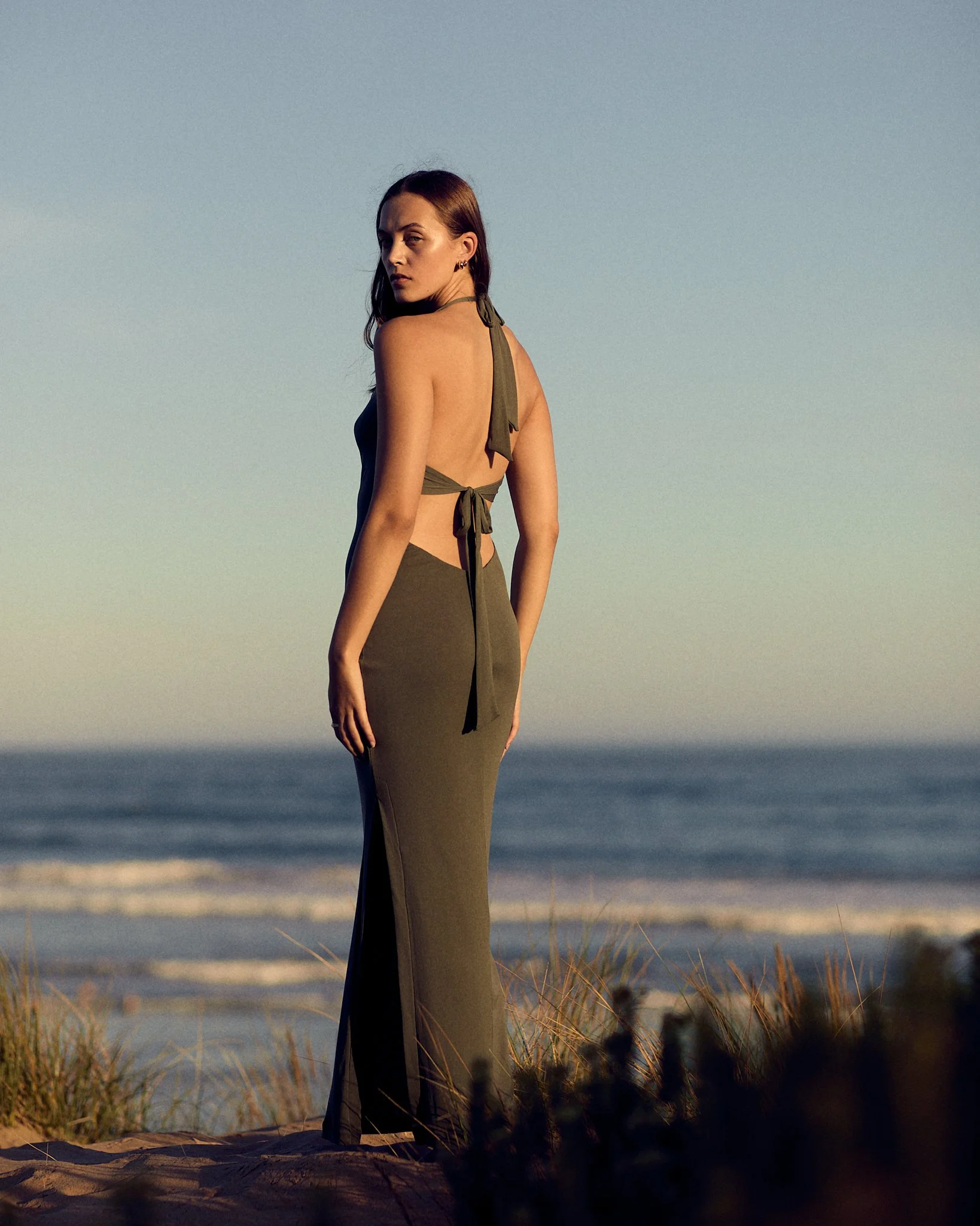 A woman in an olive green backless dress standing on a sandy beach with grass, looking over her shoulder towards the camera, with the ocean and sky in the background during sunset or late afternoon.