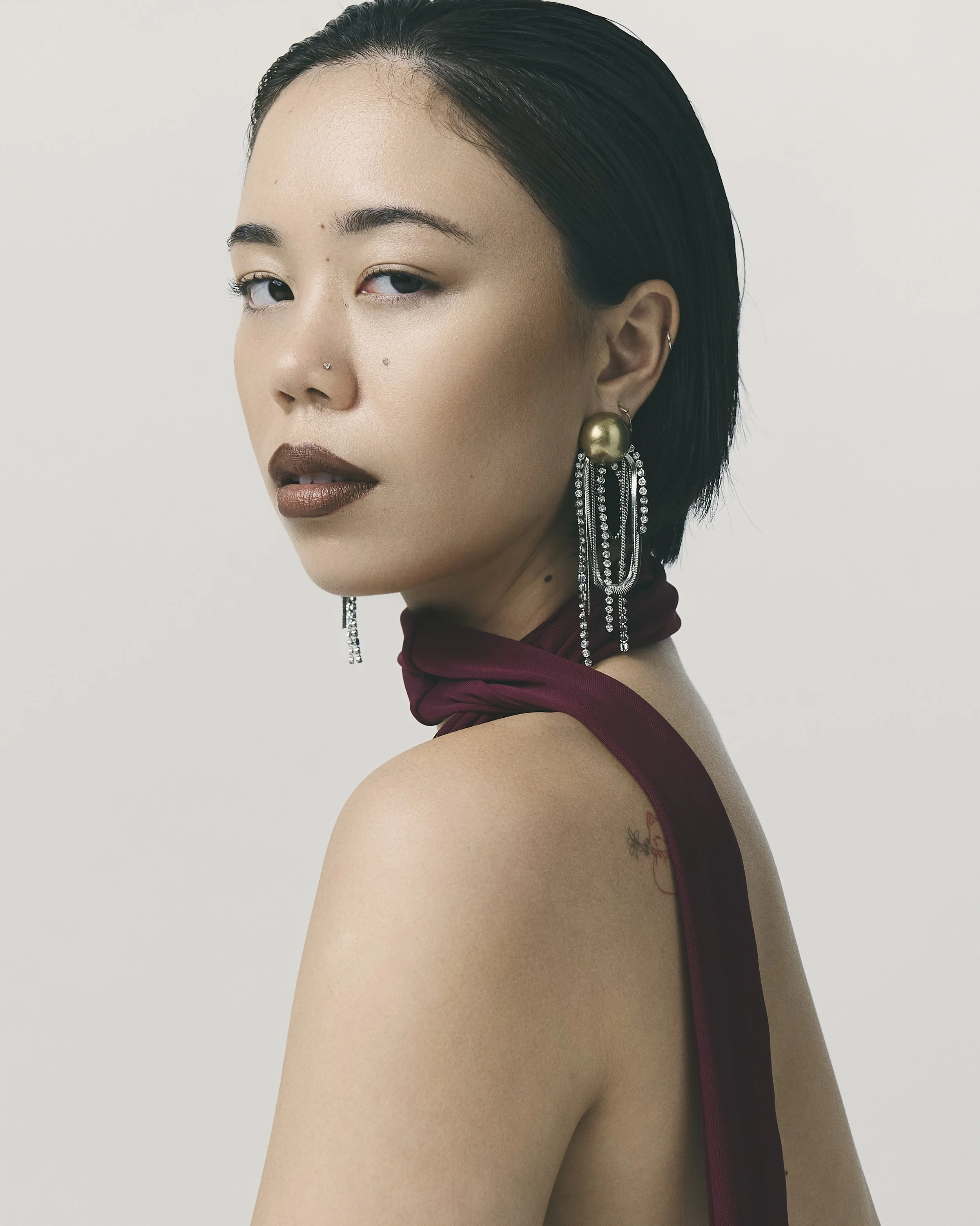A woman with short black hair, wearing large gold earrings with dangling rhinestones, a burgundy scarf around her neck, and makeup with brown lipstick.