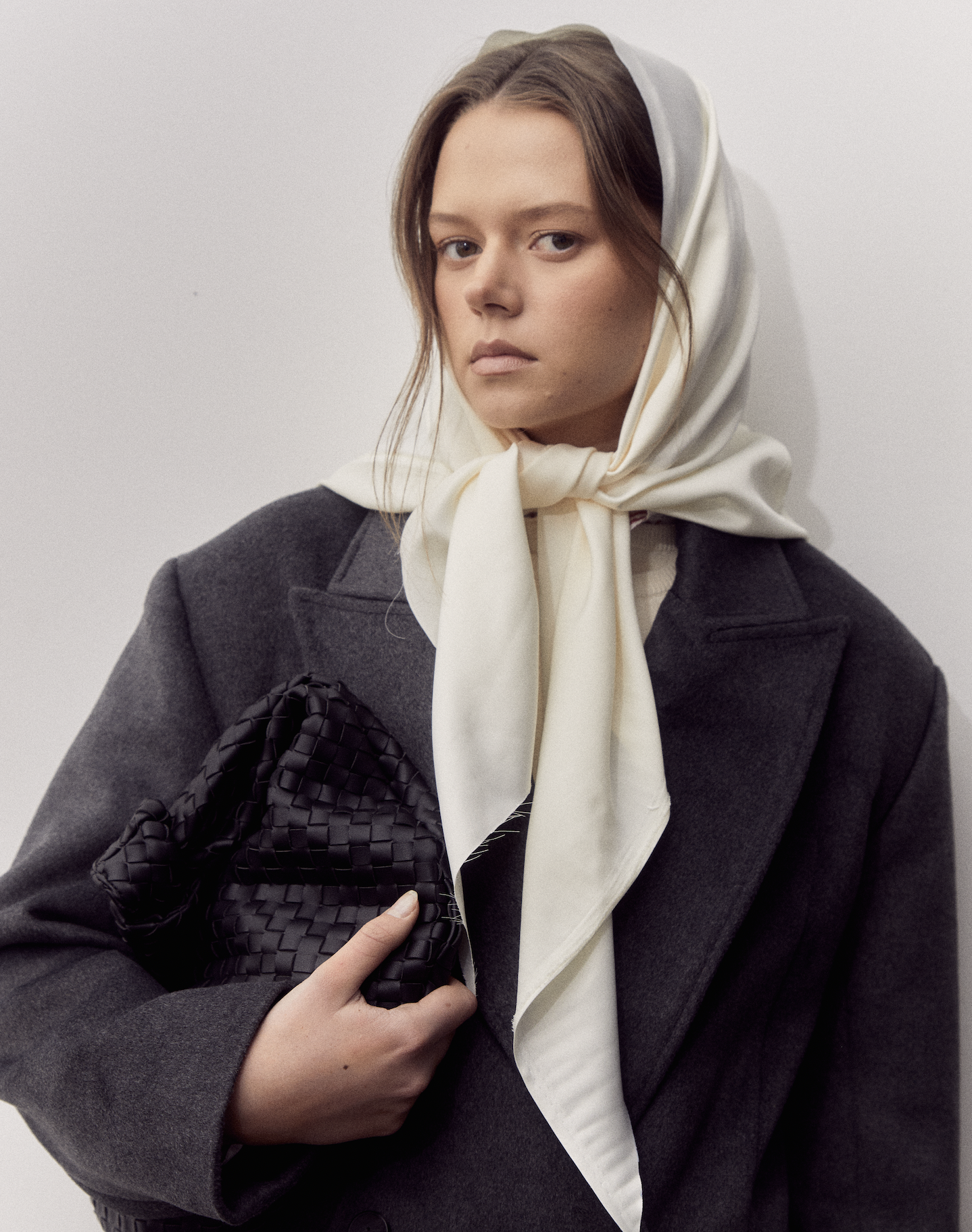 Woman with light brown hair wearing a dark gray coat, a cream-colored headscarf, and holding a black woven clutch purse, standing against a plain light background.