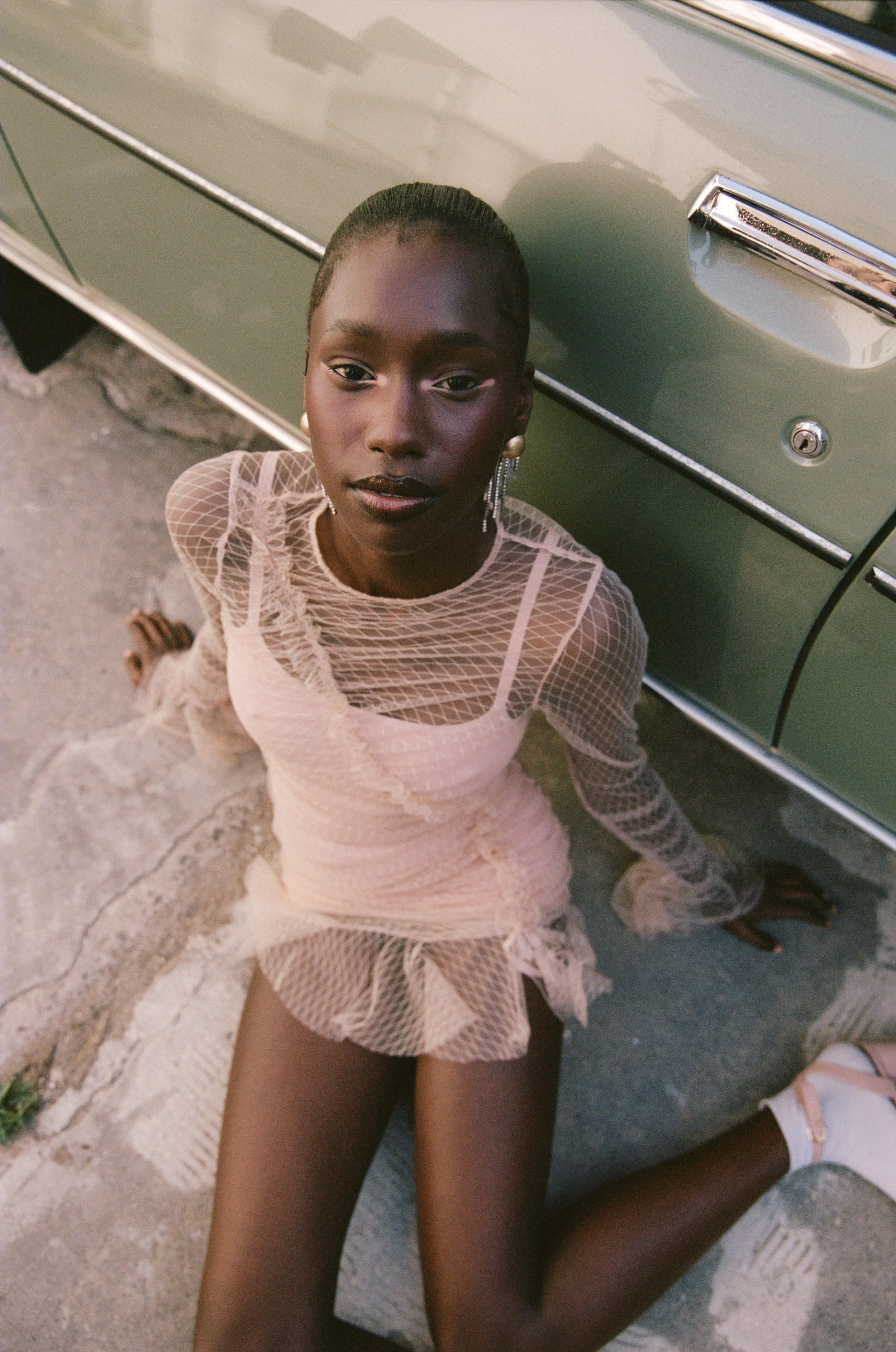 A young woman in a pink, sheer, fishnet dress with ruffled skirt and white shoes sitting on the pavement next to a vintage green car, looking up at the camera.
