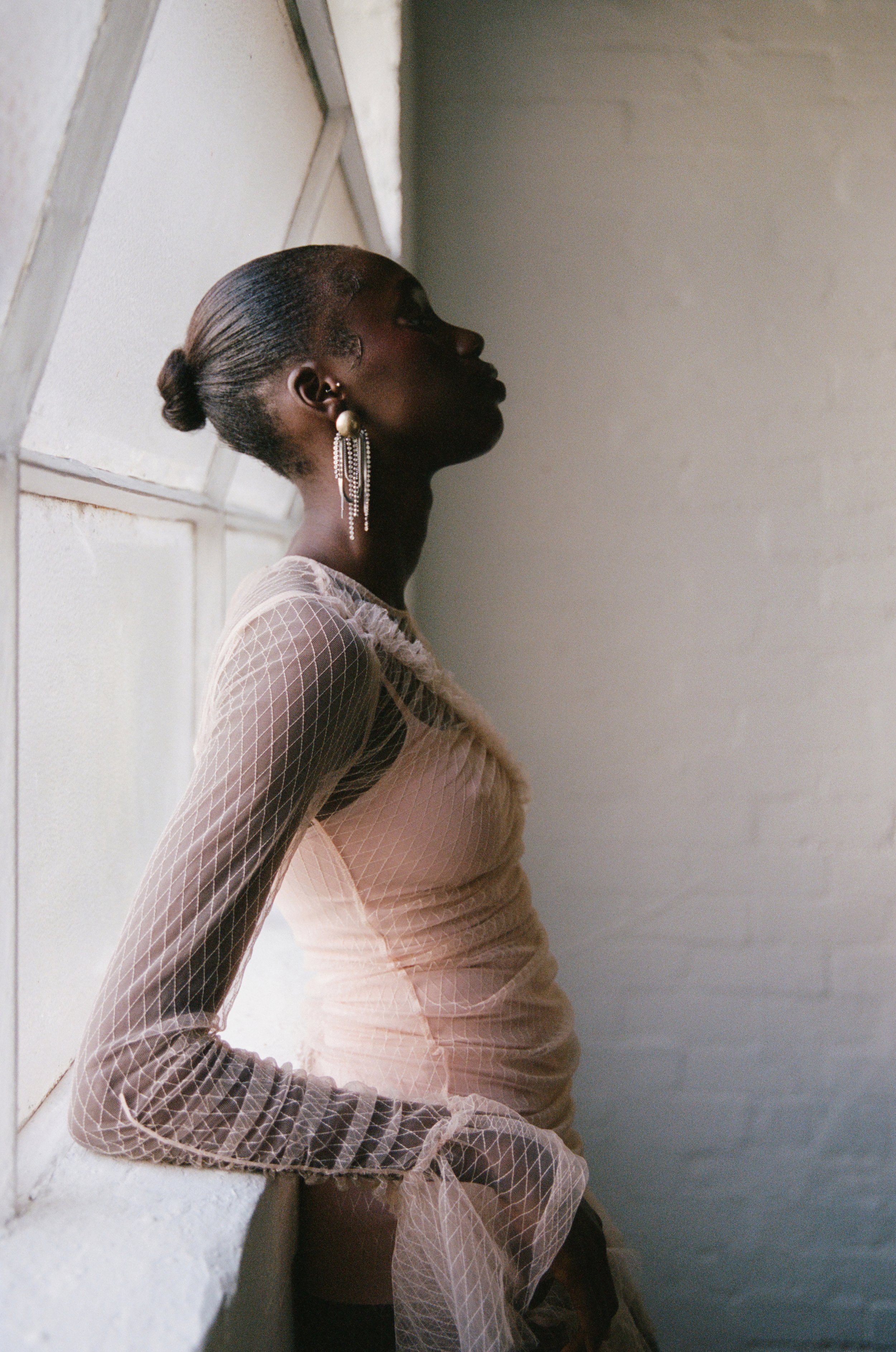 A woman with dark skin in profile stands by a window, wearing a sheer, long-sleeved top and large pearl earrings, with her hair styled in a low bun, and gazes upward.