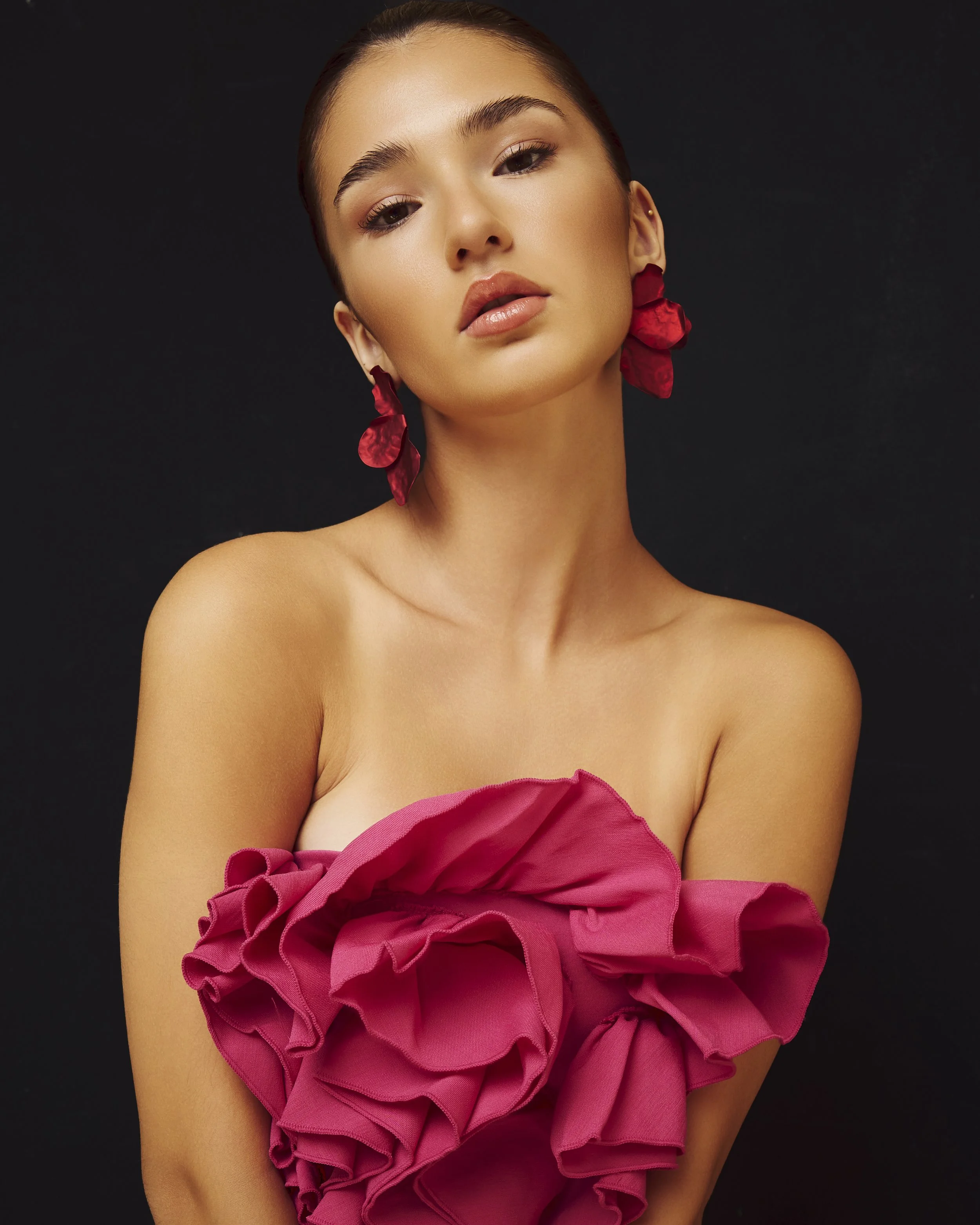 A woman with makeup, wearing red flower earrings and a pink ruffled dress, posing against a black background.