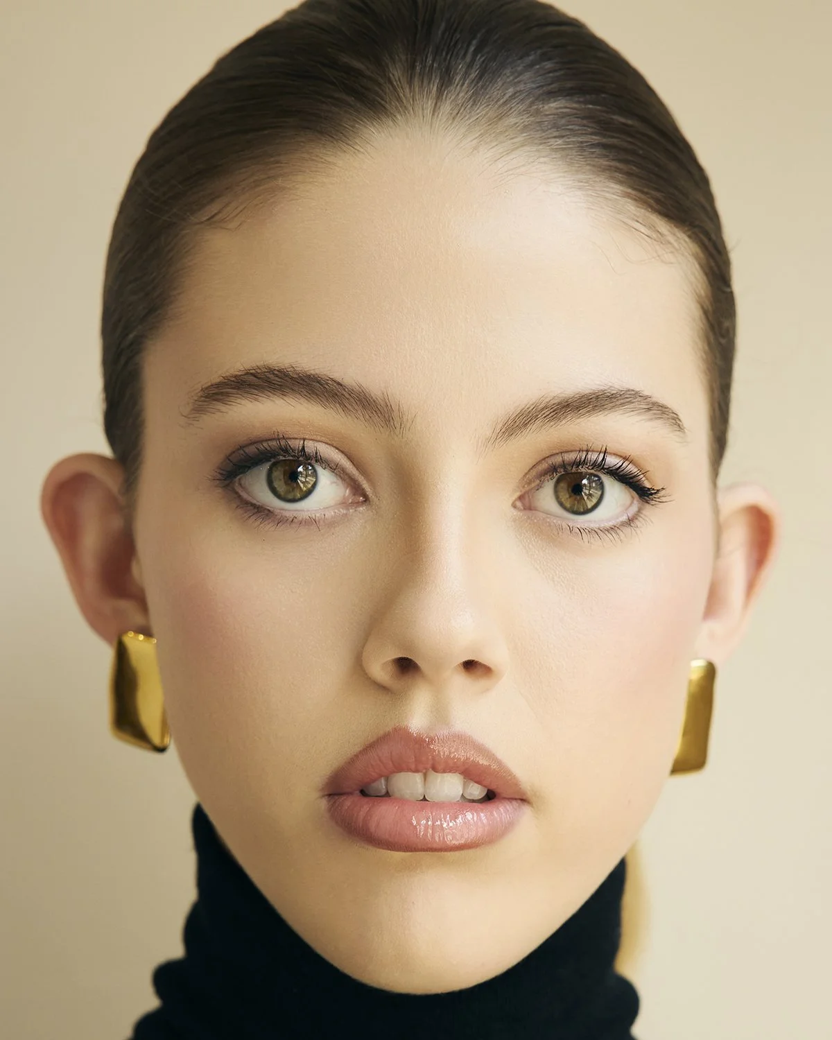 Close-up portrait of a woman with dark hair, hazel eyes, and light makeup, wearing gold earrings and a black turtleneck sweater.