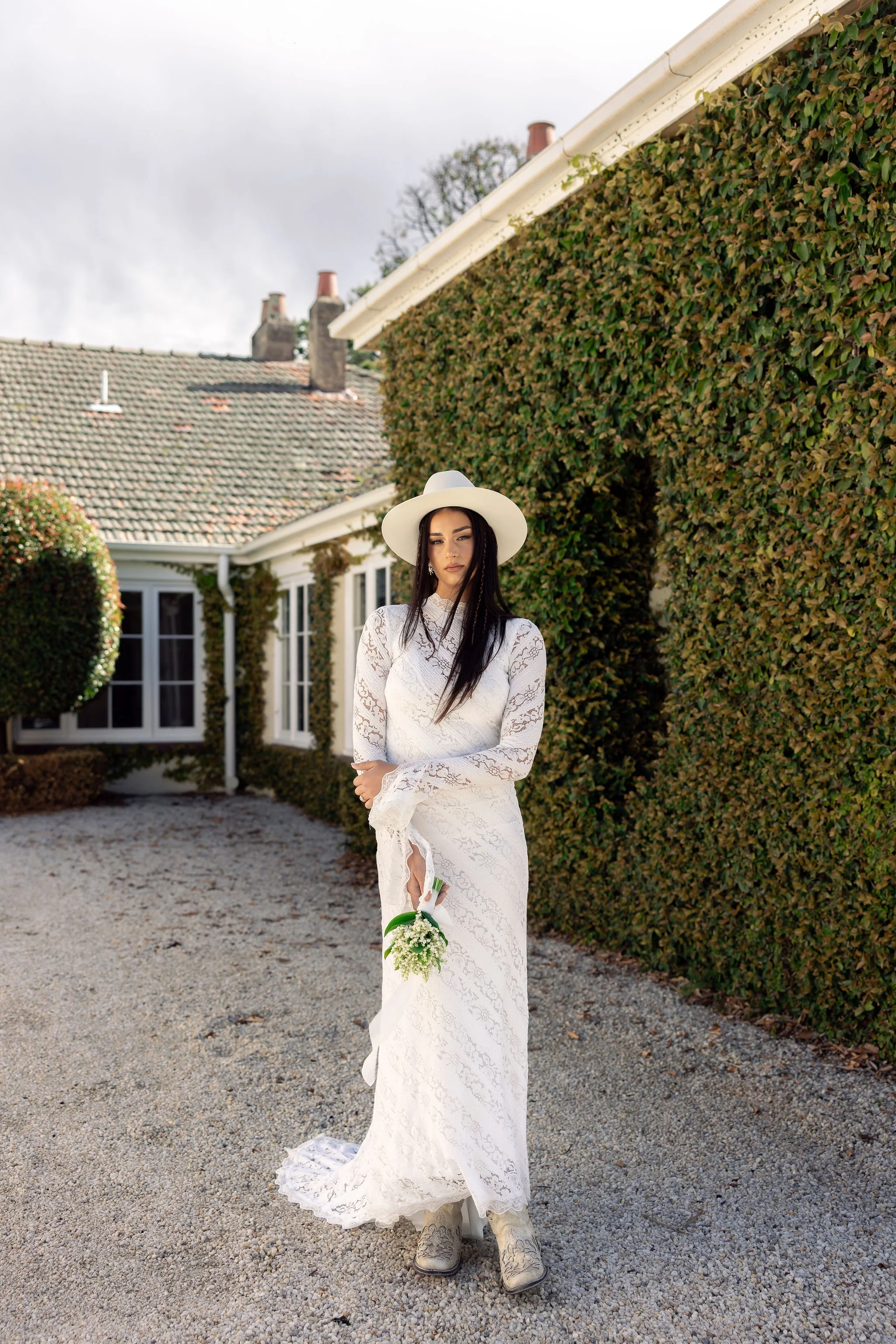 A woman in a white lace wedding dress and cowboy boots stands outdoors holding a small bouquet, wearing a wide-brimmed white hat, beside a house with green ivy on the walls and a gravel pathway.