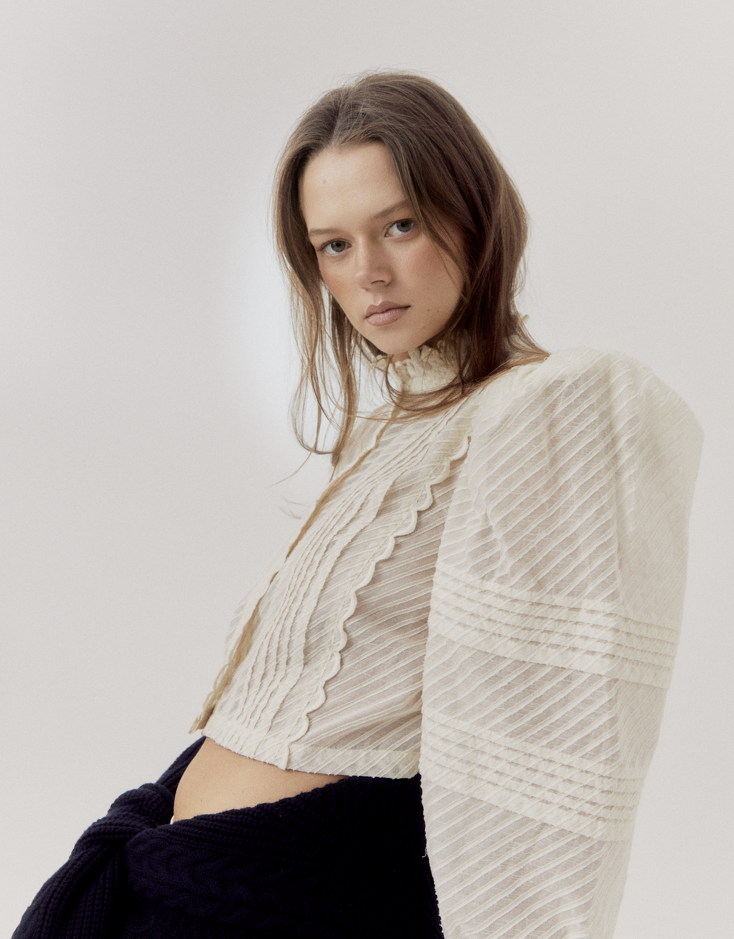 Young woman with shoulder-length brown hair wearing a cream-colored textured blouse and black pants, standing against a plain white background.