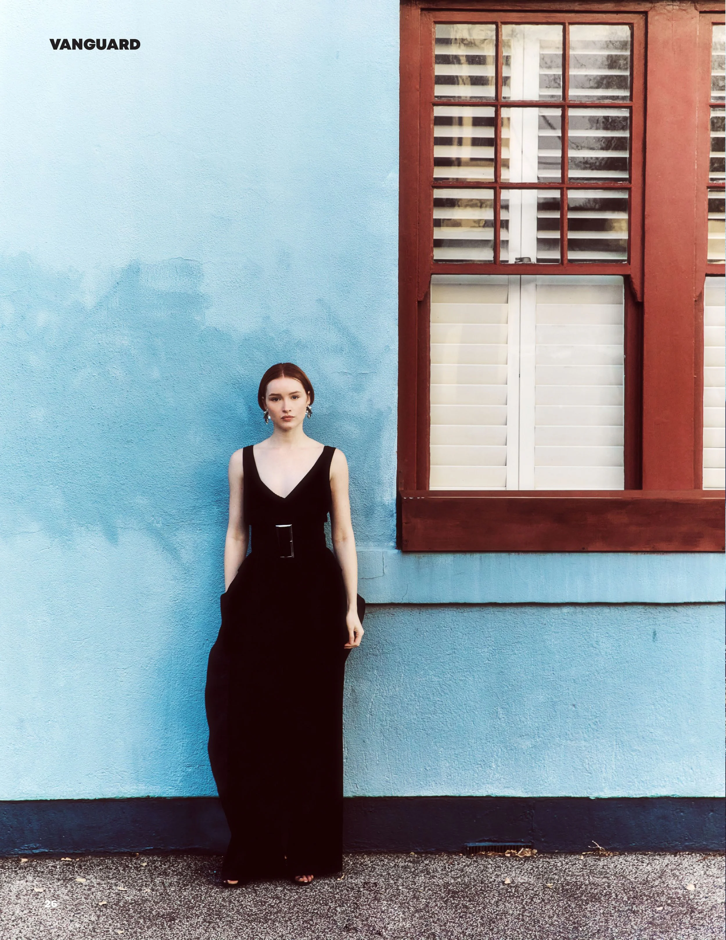 A young woman with short brown hair in a black dress standing against a blue stucco wall with a red window frame.