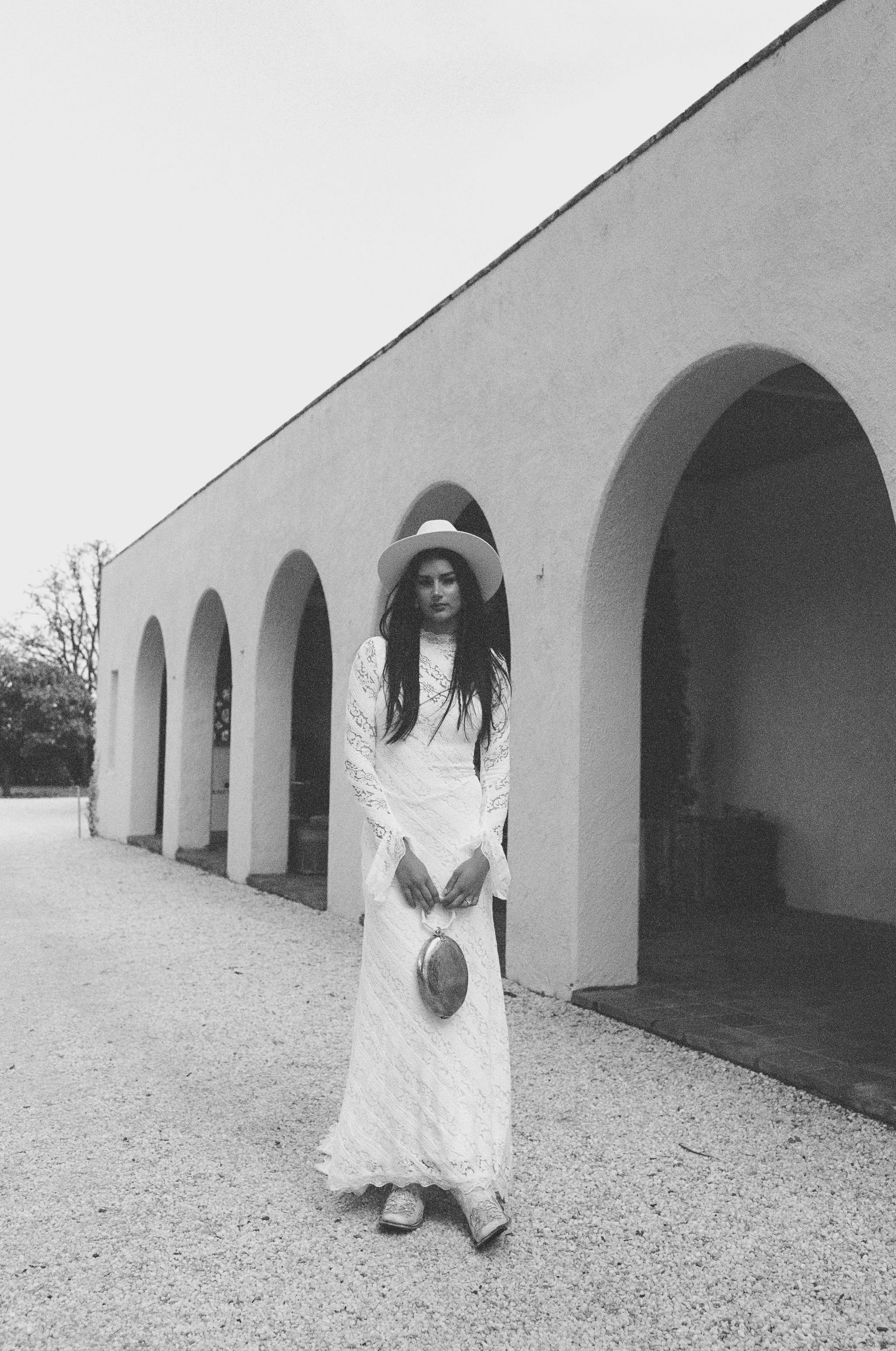 A woman in a long lace dress and hat stands outside a building with arched doorways, holding a small purse.