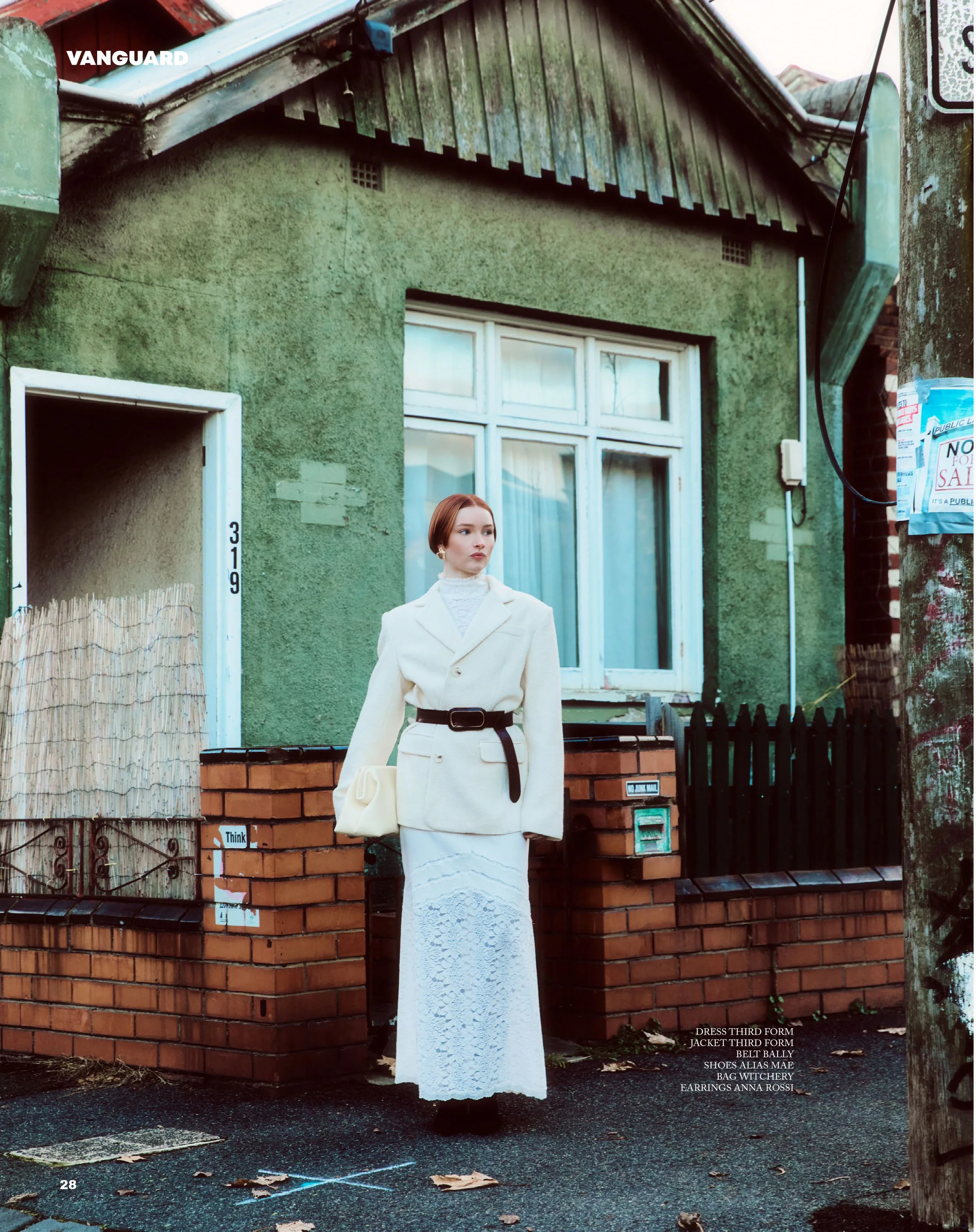 A woman dressed in a white skirt and jacket, with a black belt, standing outside a green house with a brick and black fence, and a large window, on a rainy day with fallen leaves.