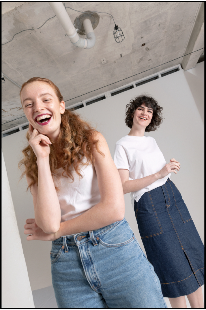 Two women with curly hair smiling and laughing in a modern room with an industrial ceiling. One woman with red hair in a white tank top and jeans stands in front, while the other with dark hair in a white t-shirt and denim skirt stands behind.