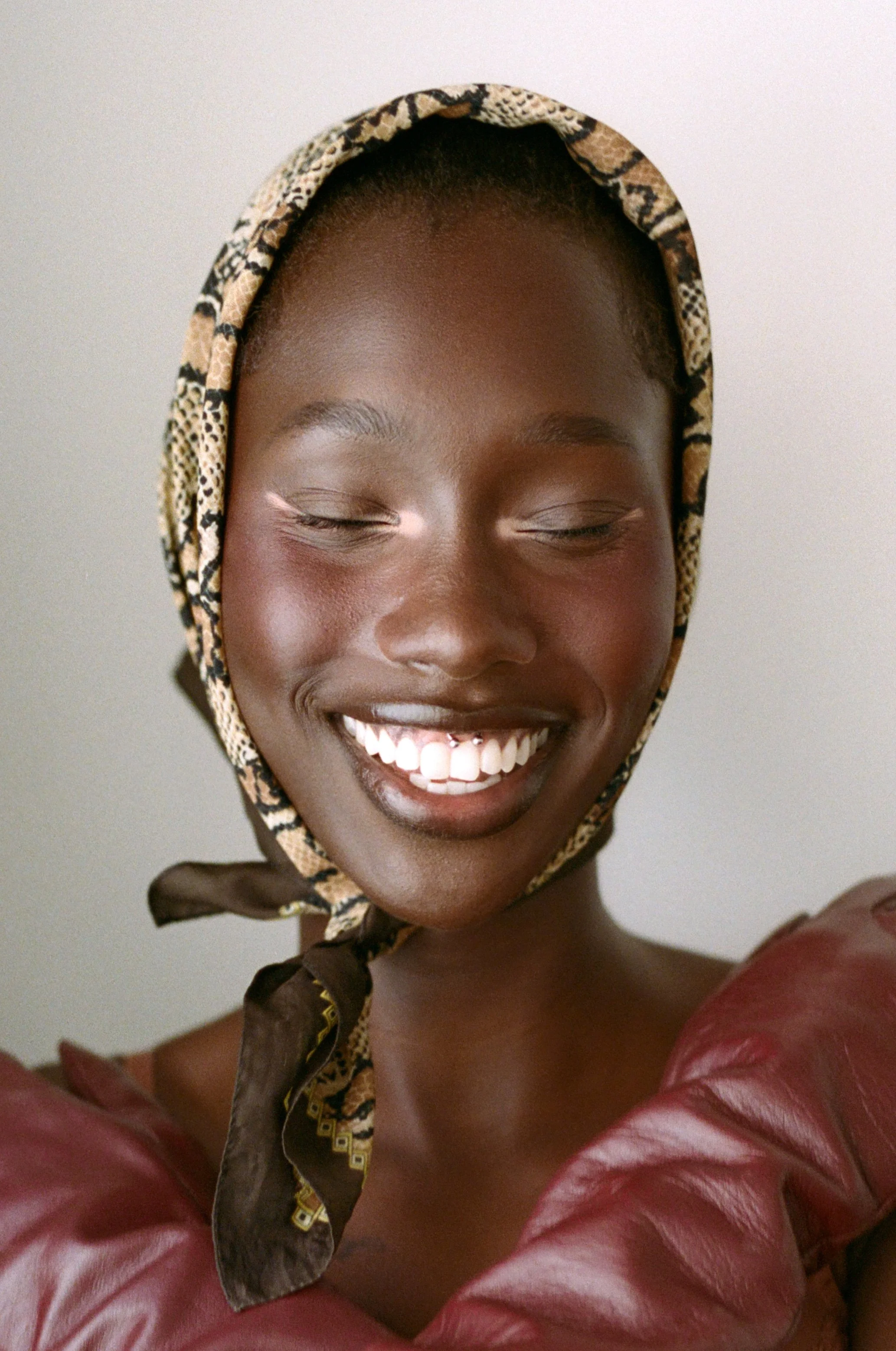 A smiling woman with dark skin and closed eyes, wearing a patterned headscarf and a red leather top.