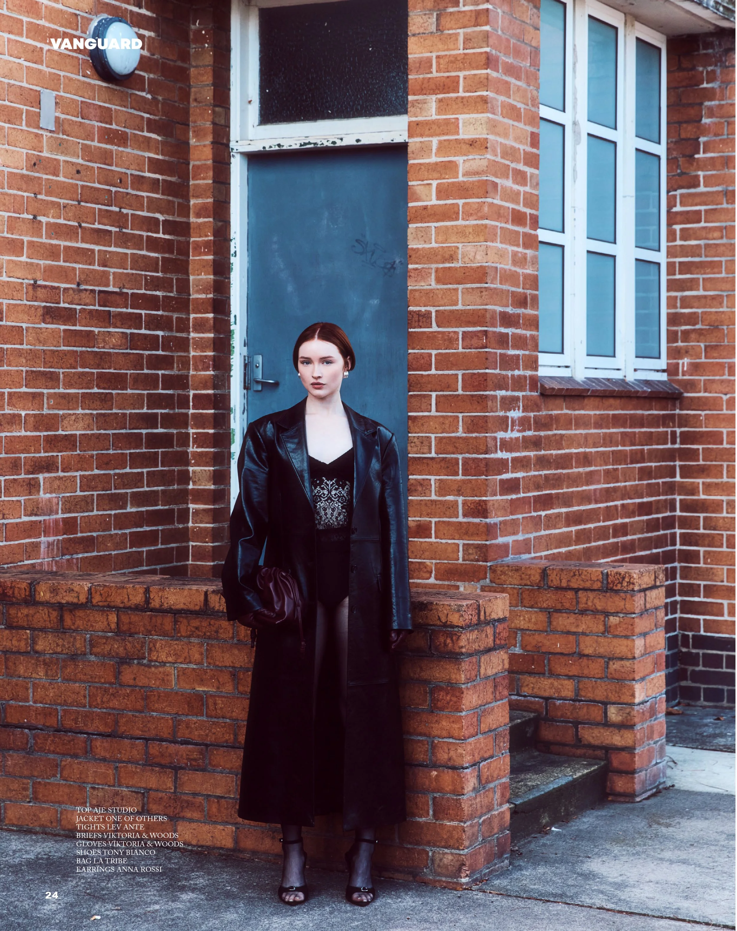A woman with red hair stands outside a brick building, wearing a black leather coat, lace top, long skirt, and high heels, holding a burgundy clutch purse.