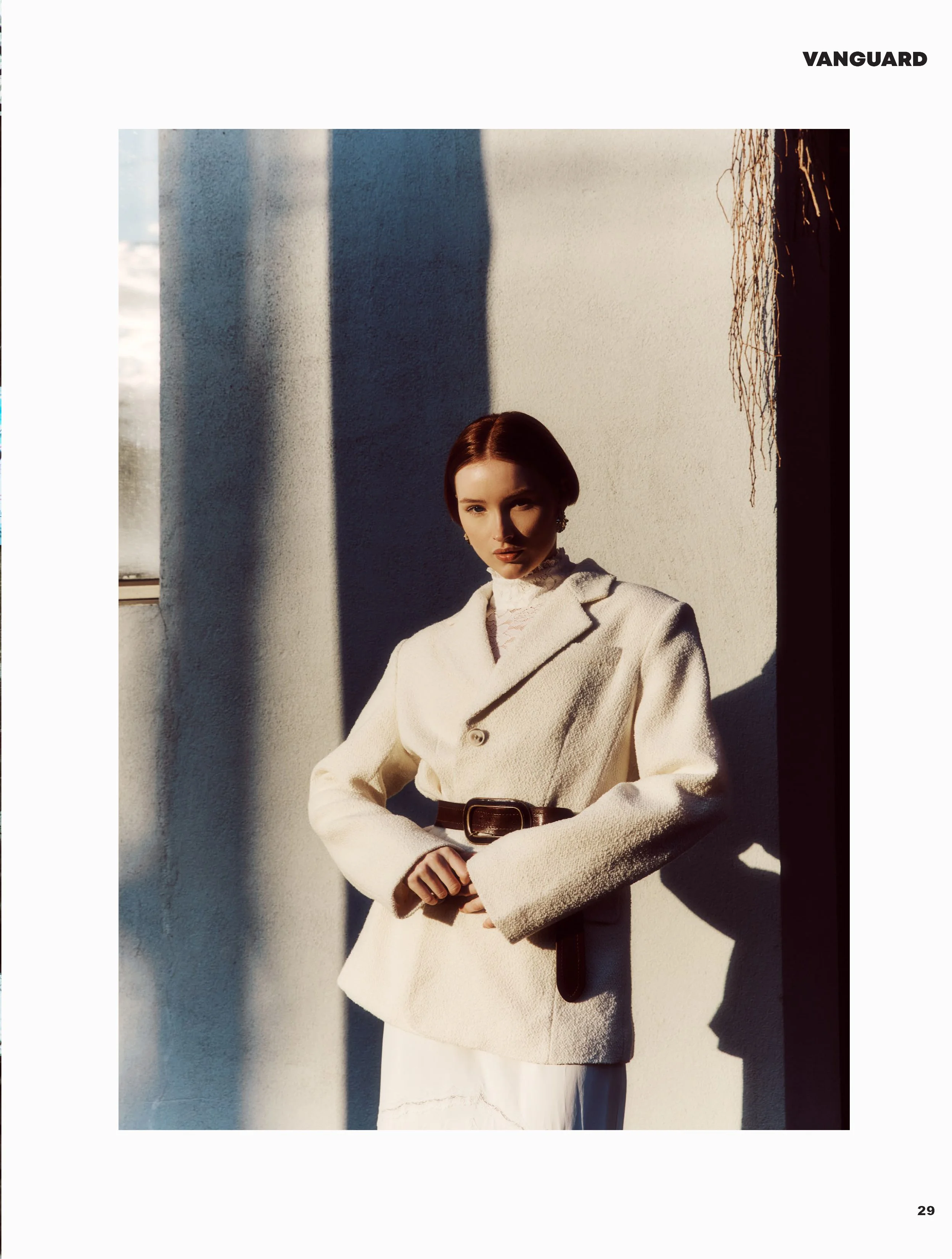 Woman with short brown hair wearing a white textured blazer and a black belt, standing against a white wall with shadows and sunlight