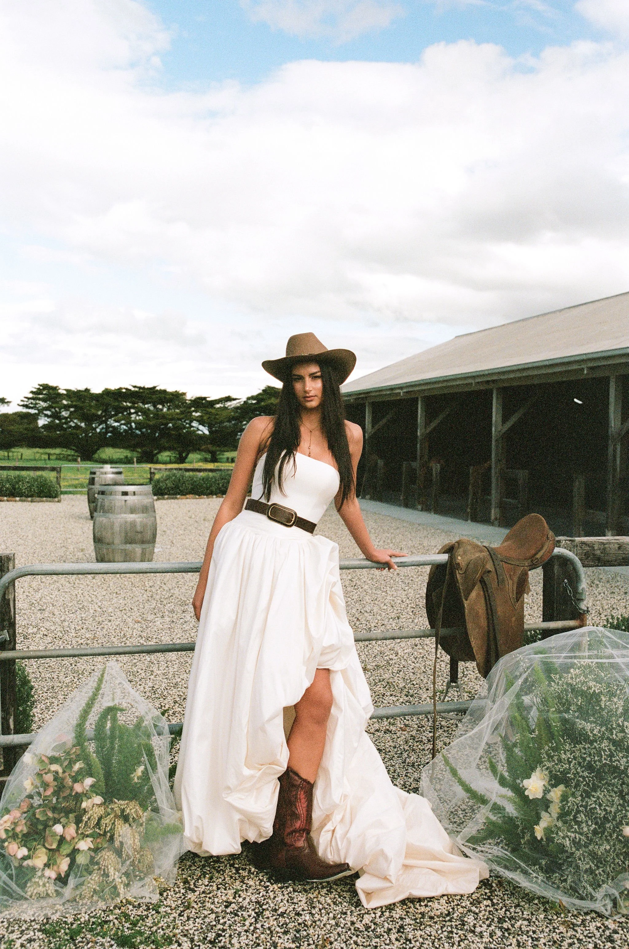 A woman dressed in a white strapless dress, brown cowboy hat, and boots standing outdoors near a fence, with flower arrangements around her.