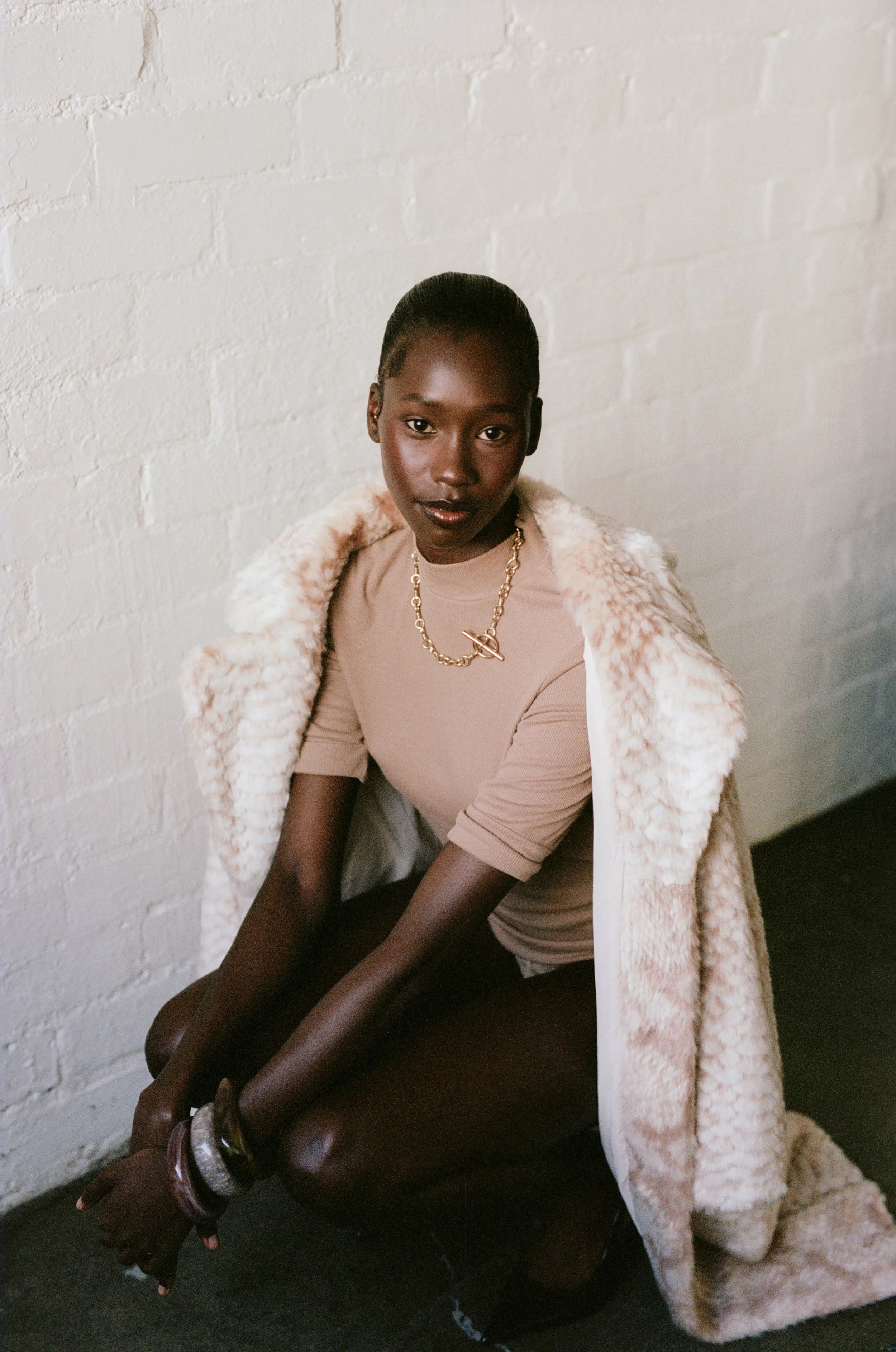 A woman with dark skin and hair pulled back, sitting on the floor against a white brick wall. She is wearing a beige top, a faux fur coat draped over her shoulders, a gold necklace, and bracelets, looking directly at the camera with a confident expre