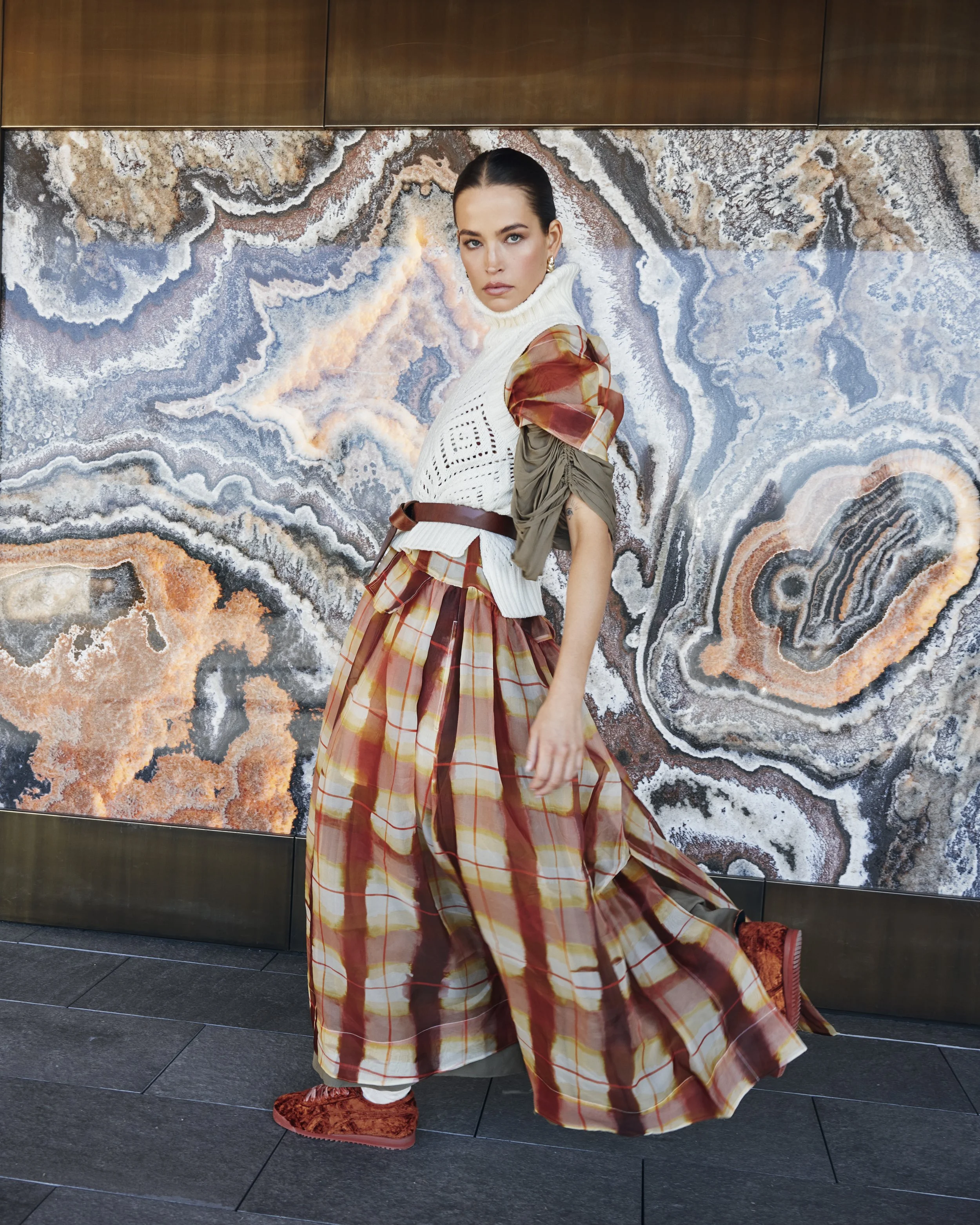 A woman in fashion-forward clothing standing in front of a colorful abstract stone wall. She wears a white turtleneck sweater vest, a plaid skirt, and layered sleeves with mixed fabrics. She has dark hair pulled back and a serious expression.