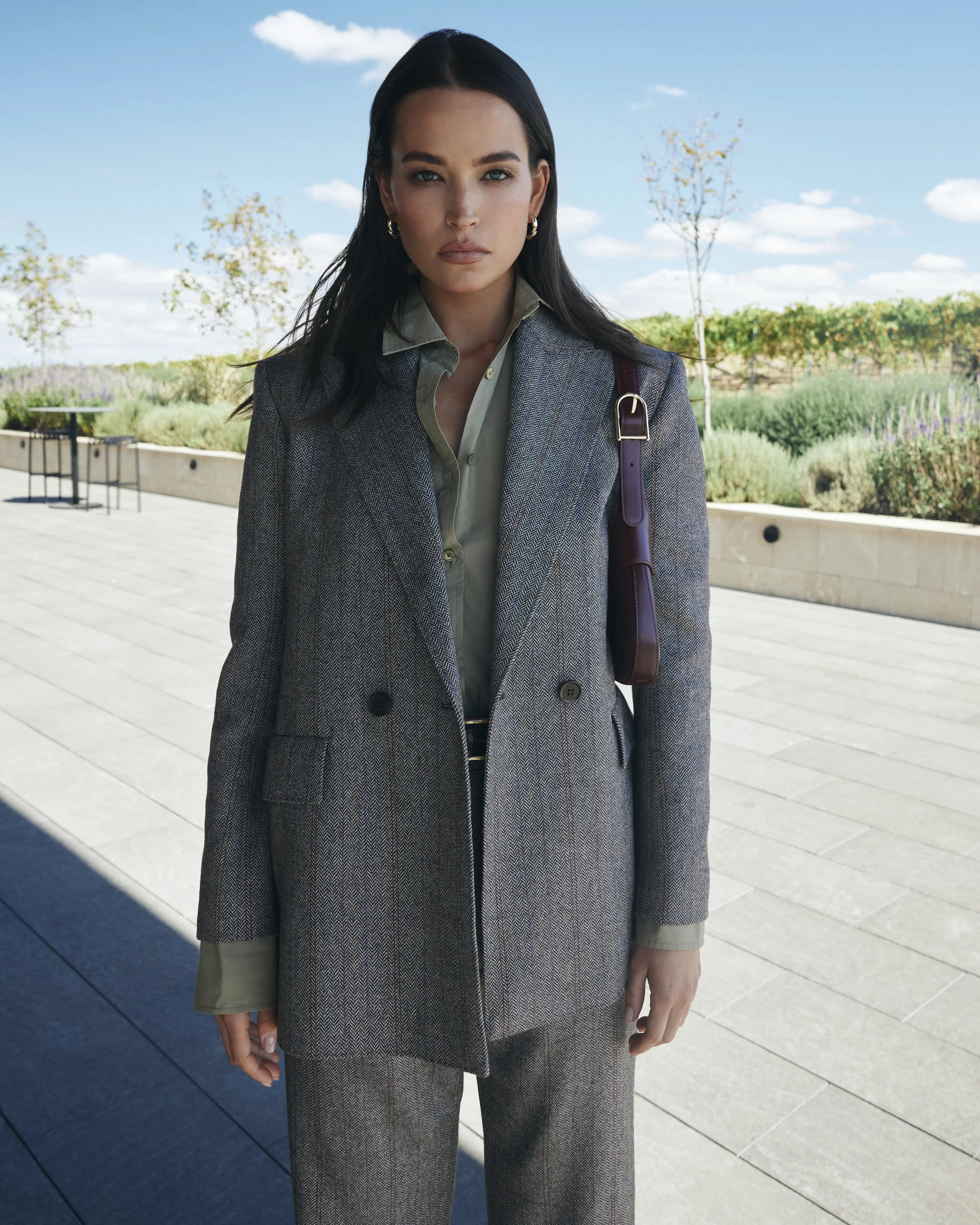 A woman with dark hair standing outdoors on a sunny day in a gray plaid blazer, matching pants, light green shirt, and a dark shoulder bag, with a background of trees and sky.