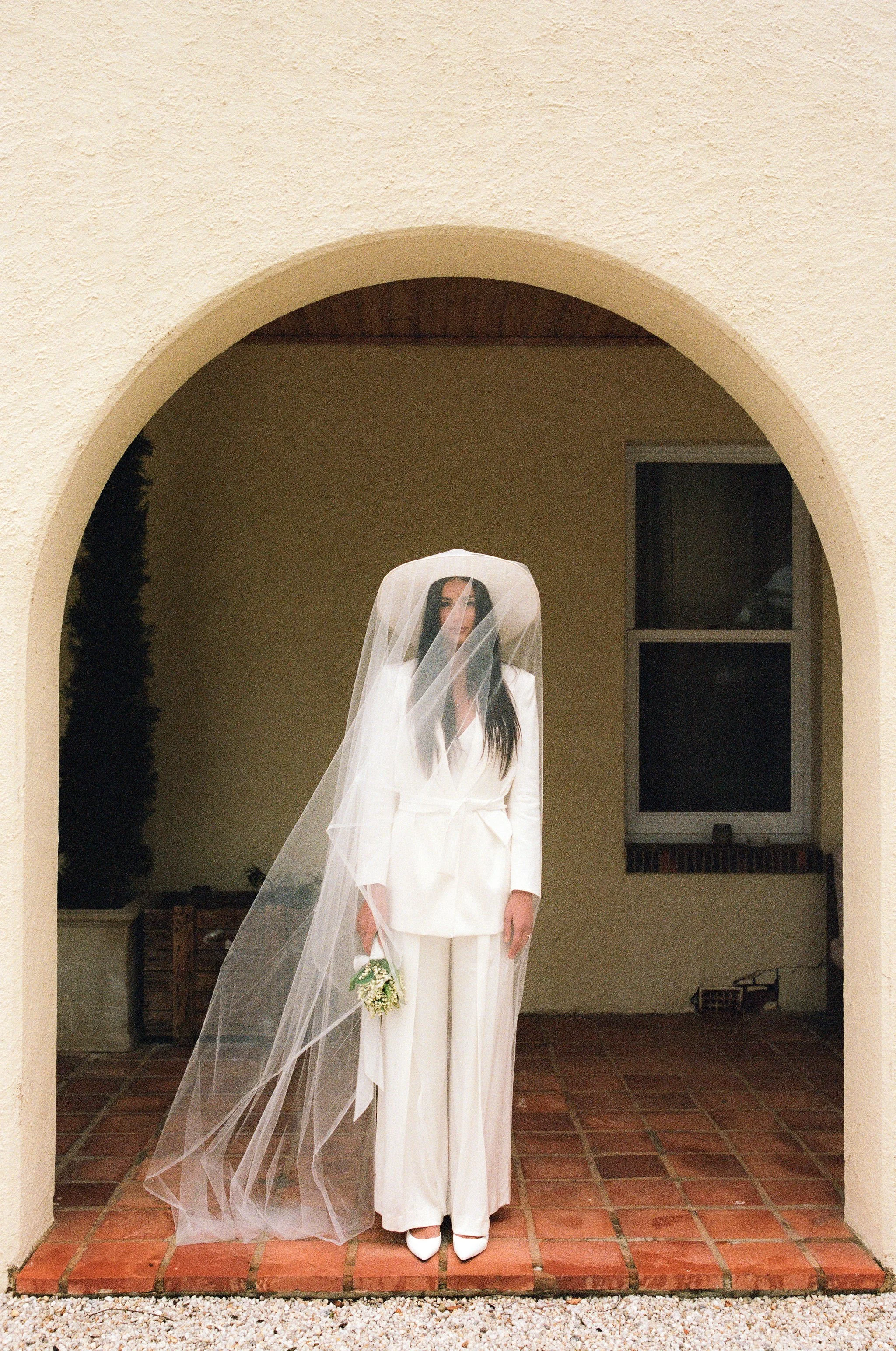 Brunette woman in a white bridal suit with a veil and holding a small bouquet, standing under an archway with a yellow wall and a window in the background.