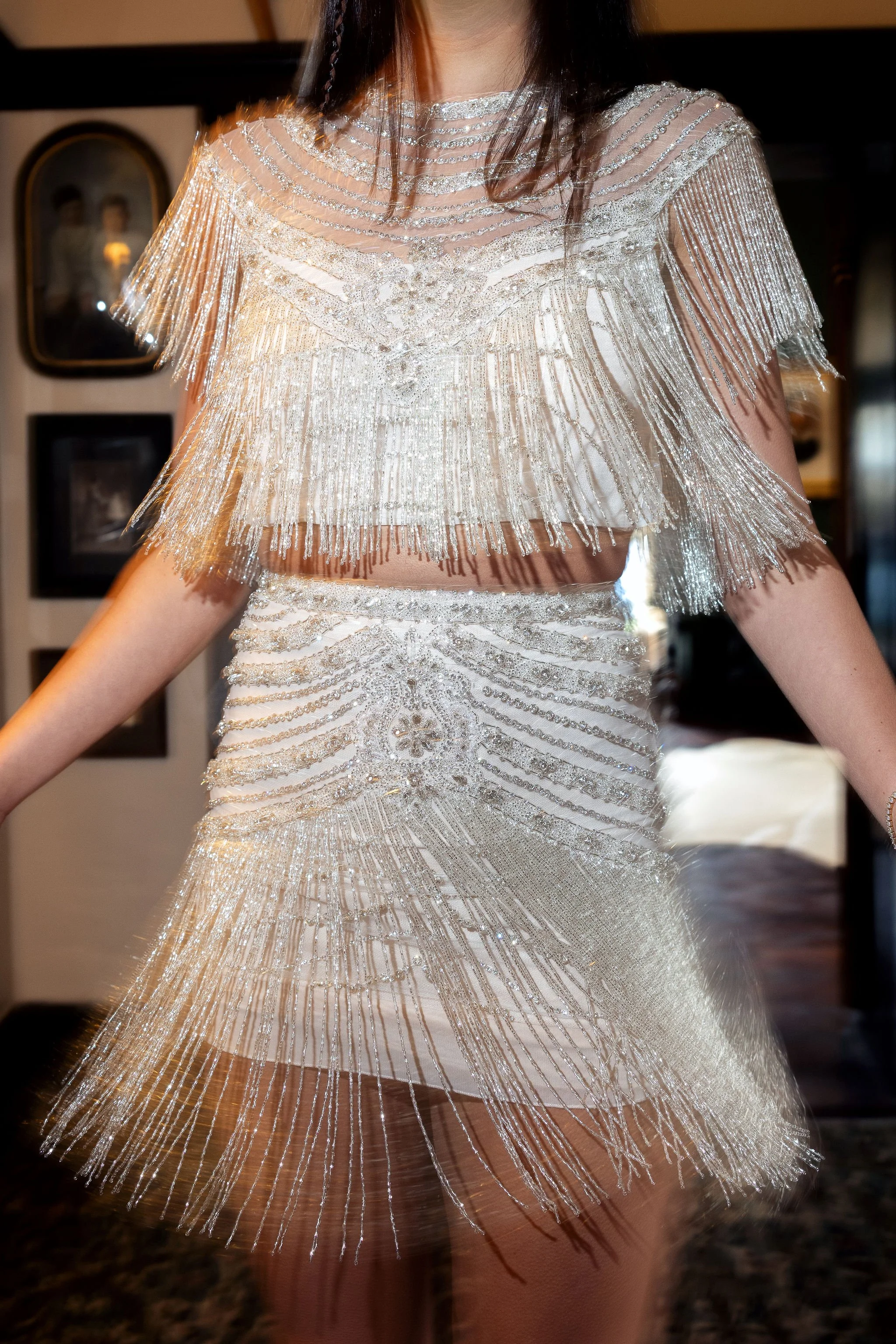 A woman is wearing an ornate, silver beaded two-piece outfit featuring a fringe top and matching skirt, both heavily decorated with sequins and beads.