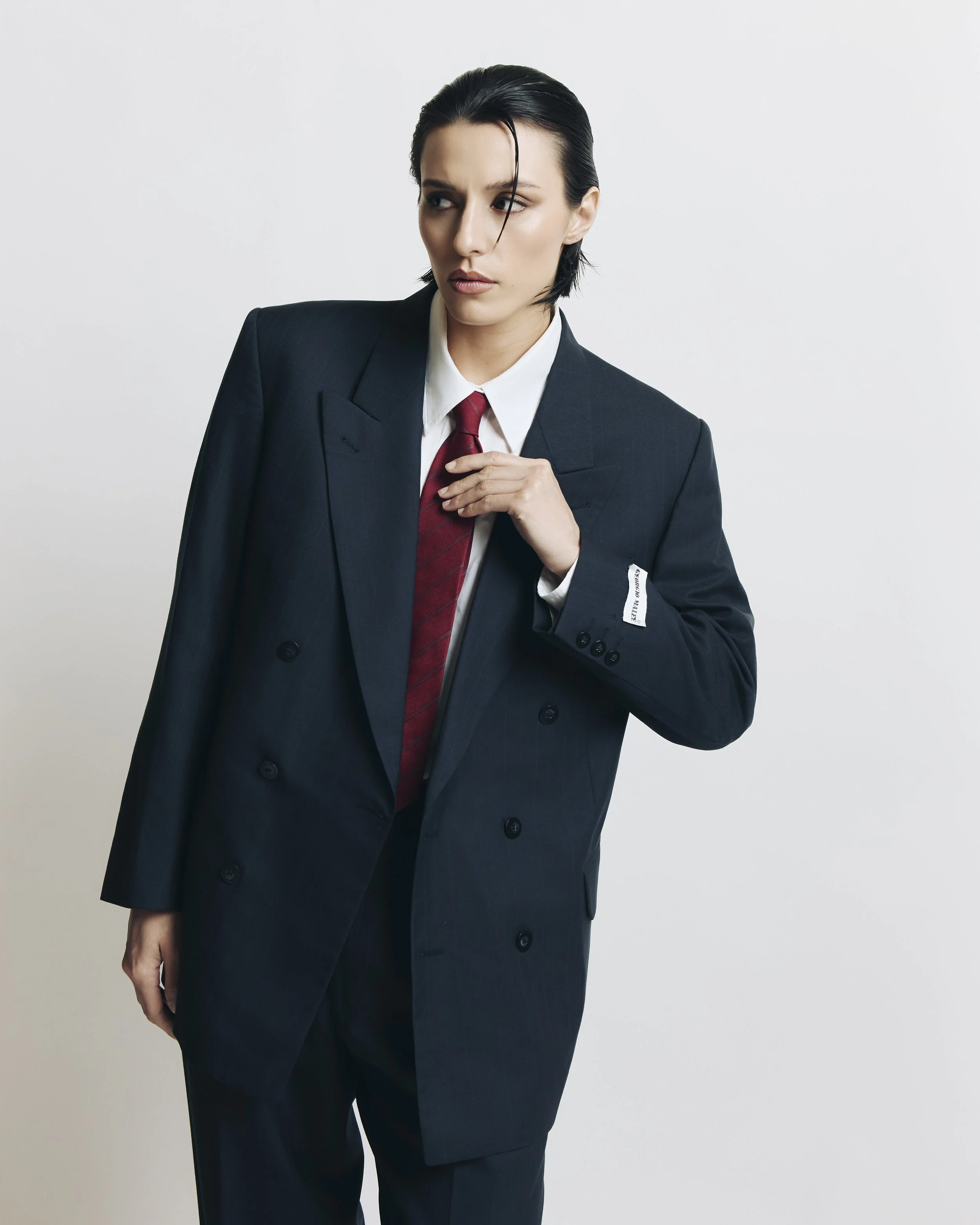Woman in a dark suit with a red tie, standing against a plain white background, looking to the side with her left hand adjusting her collar.