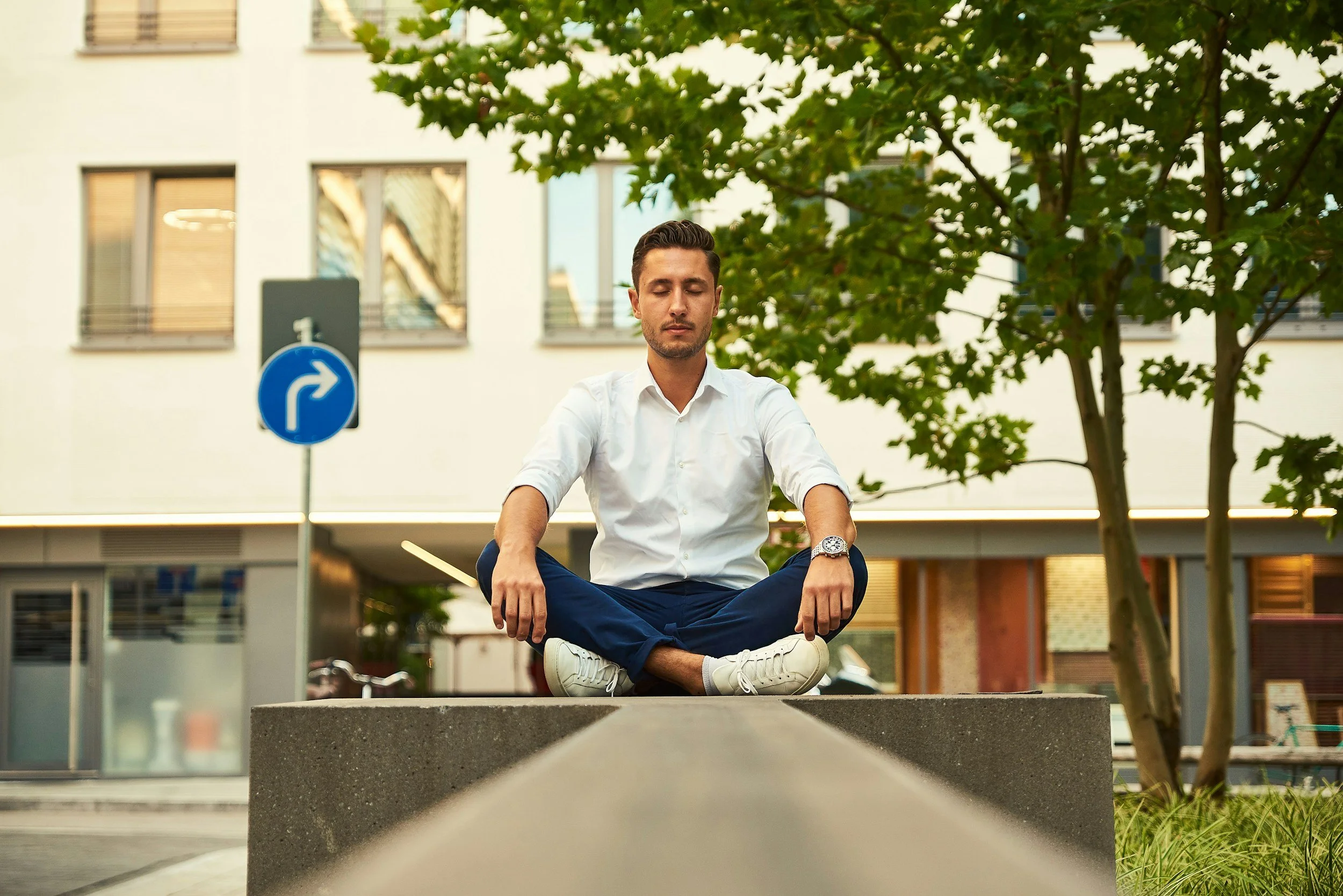 Man sitting cross legged outdoors with eyes closed, practicing meditation near trees and city buildings.