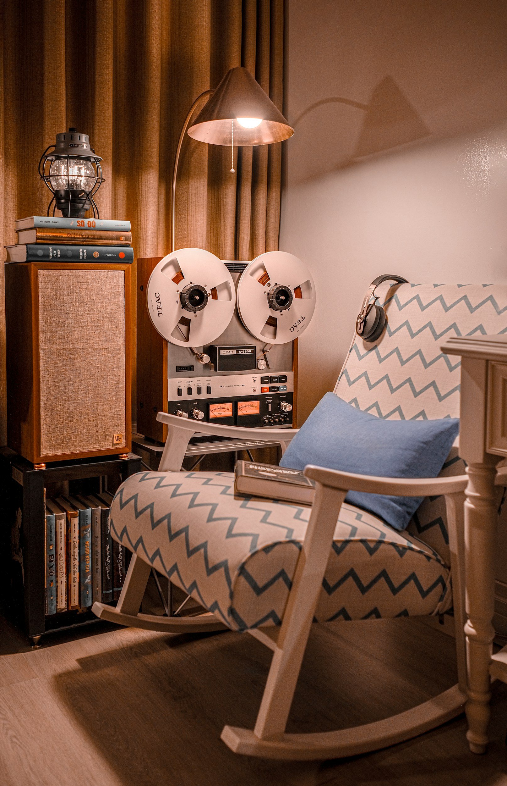 A cozy corner with a patterned rocking chair, a blue pillow, a book, a vintage reel-to-reel tape recorder, a wooden speaker with a woven front, a lamp, a lantern, stacked books, and a bookshelf with more books.