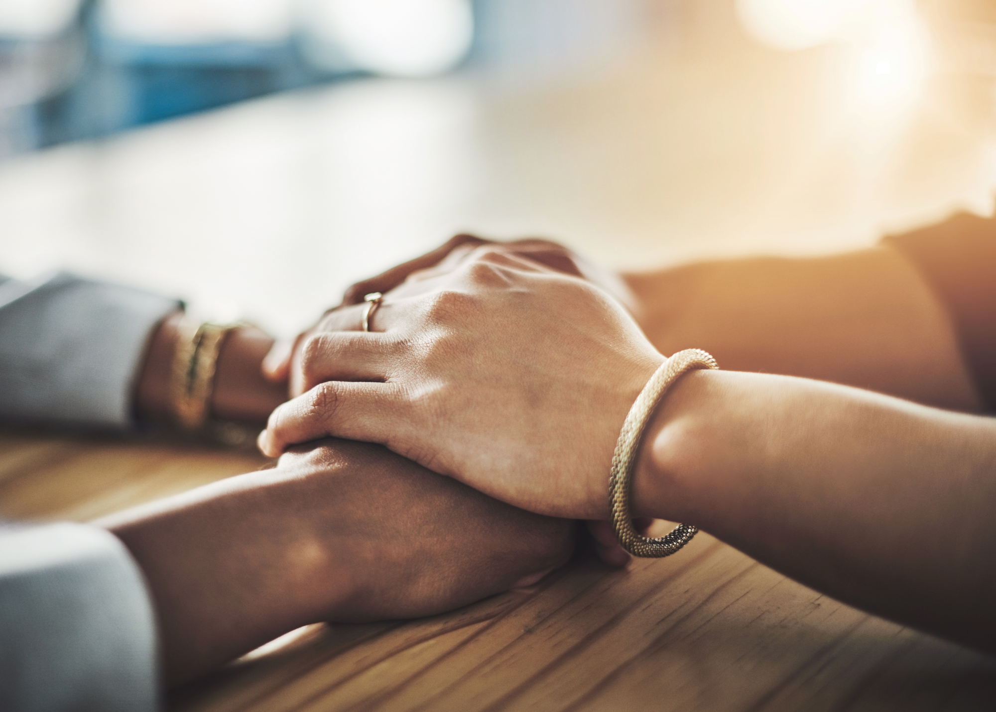 Two sets of hands rest together on a wooden table in warm light, one wearing a bracelet, suggesting a moment of quiet connection.