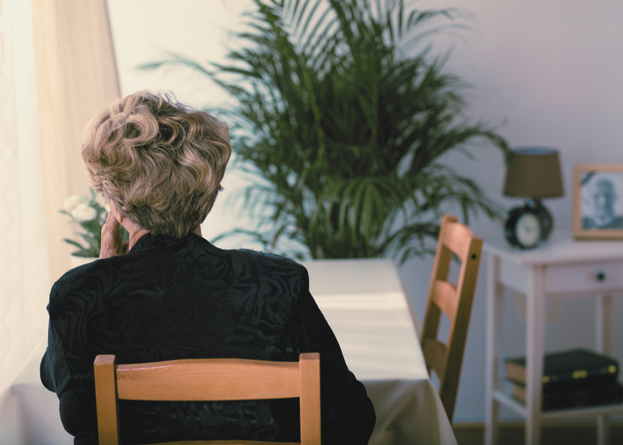 An older woman sits alone in a quiet room, her back to the camera, looking toward a window. A chair and nightstand are visible beside her.